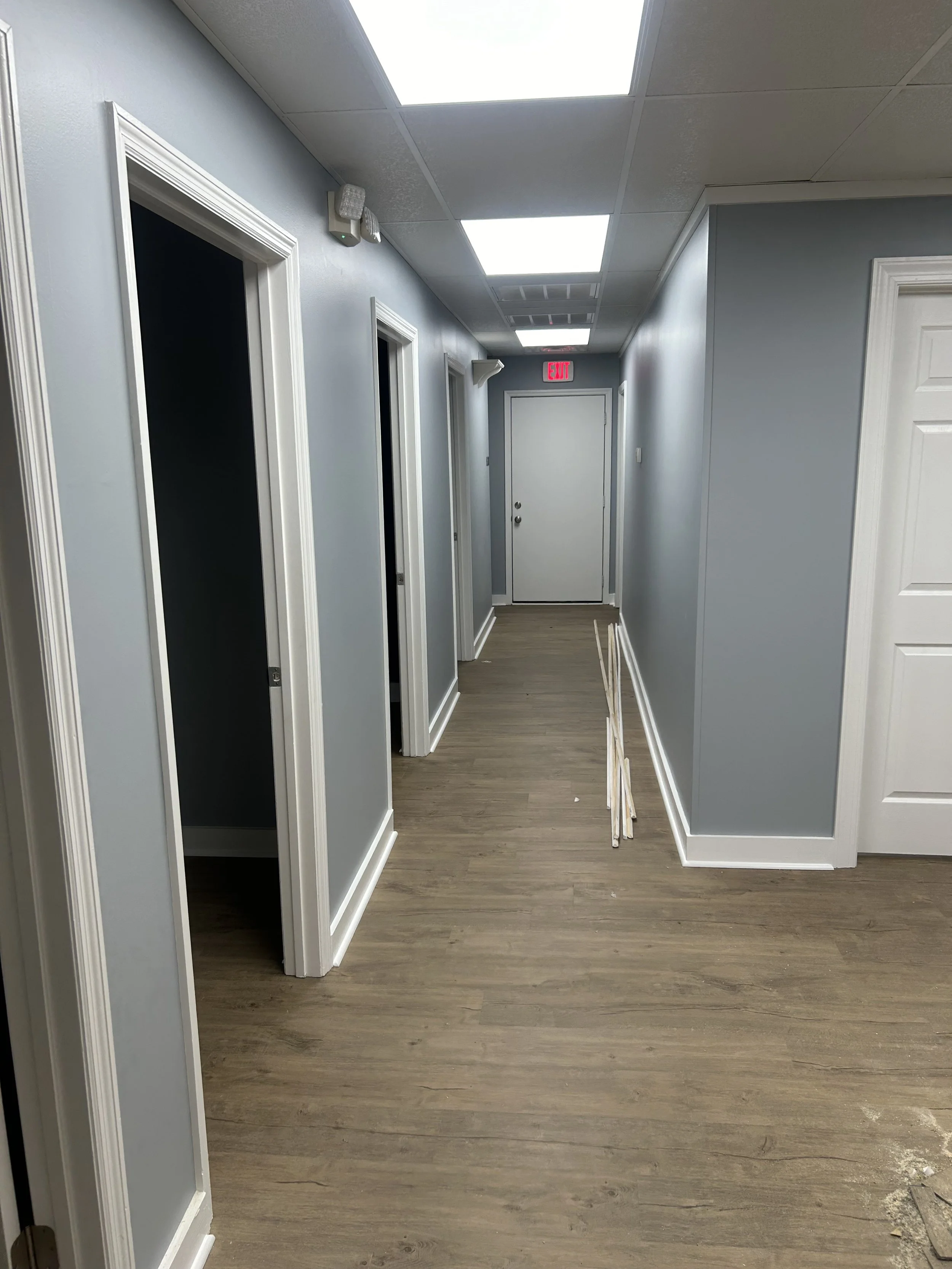 Empty office hallway with light blue walls, white doorways, wooden flooring, and a stack of white trim or baseboards leaning against the wall.