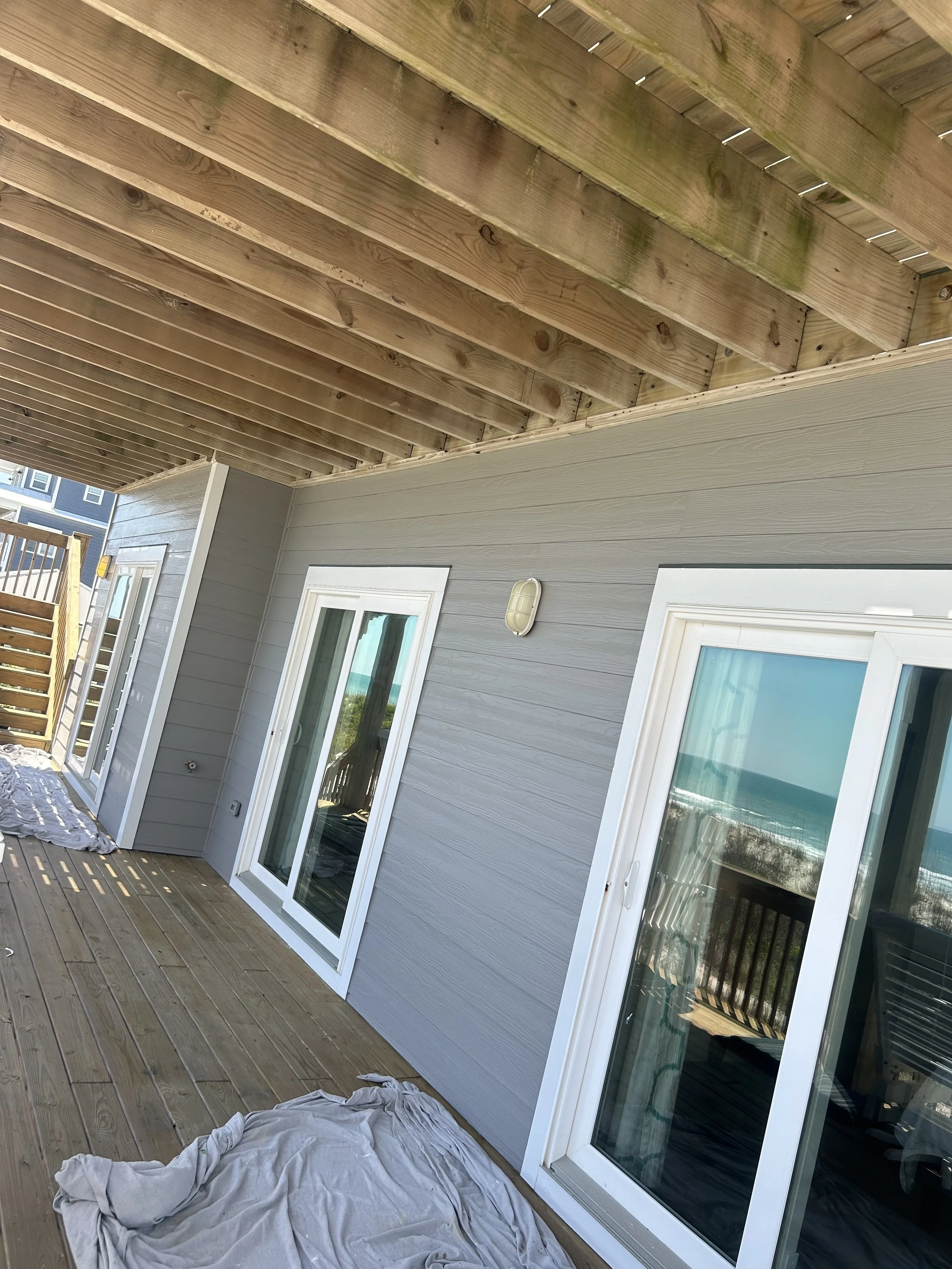 View of a wooden deck with sliding glass doors leading to the interior, overlooking the ocean in the distance, with construction materials and towels on the deck.