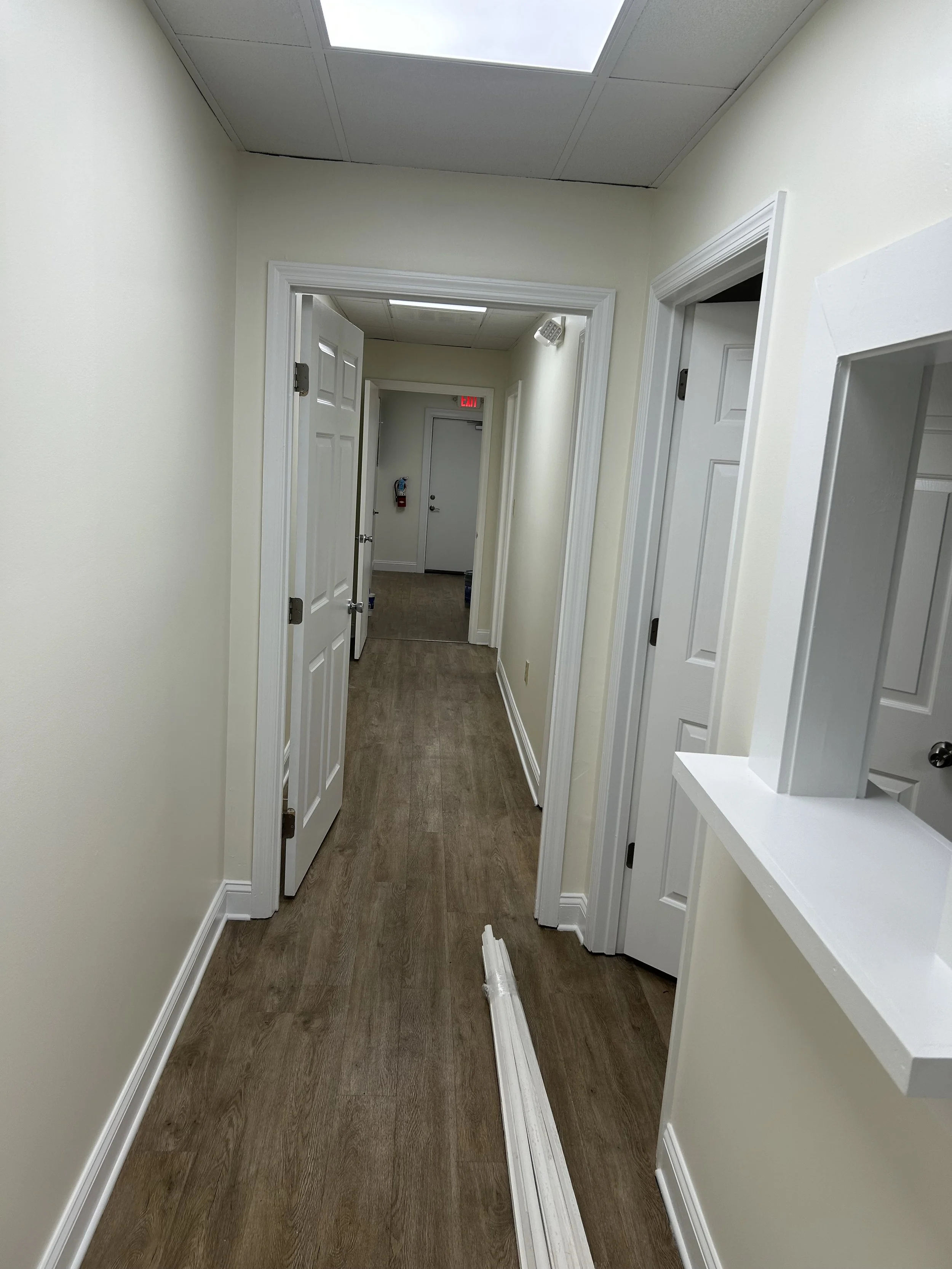 Empty hallway with light yellow walls, white trim, brown wood floor, and ceiling lights, leading to a door at the end.