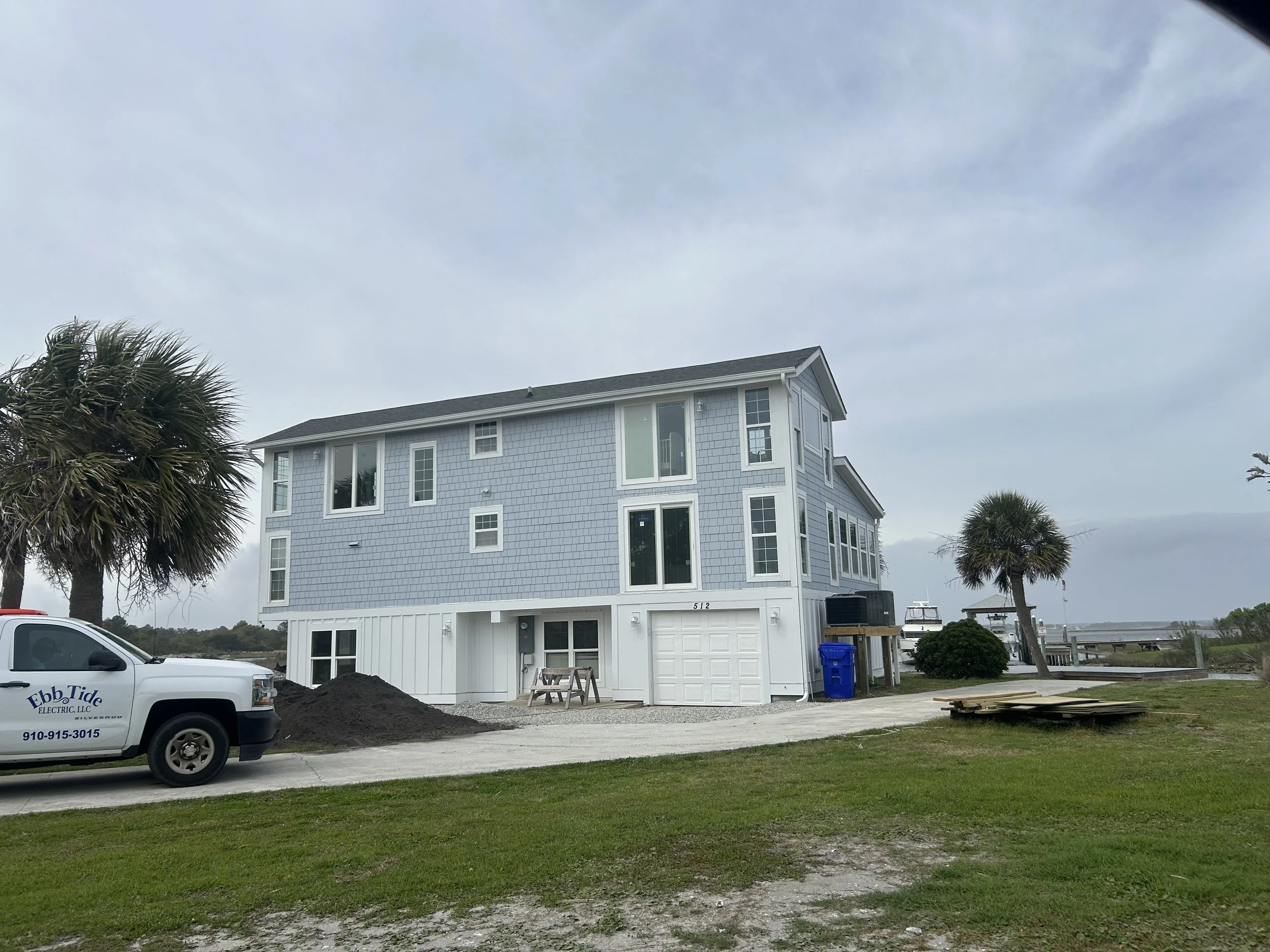 A multi-story blue house with several windows, near a waterfront, with palm trees, construction materials, and a white truck labeled 'Ebb Tide Electric LLC' parked nearby.
