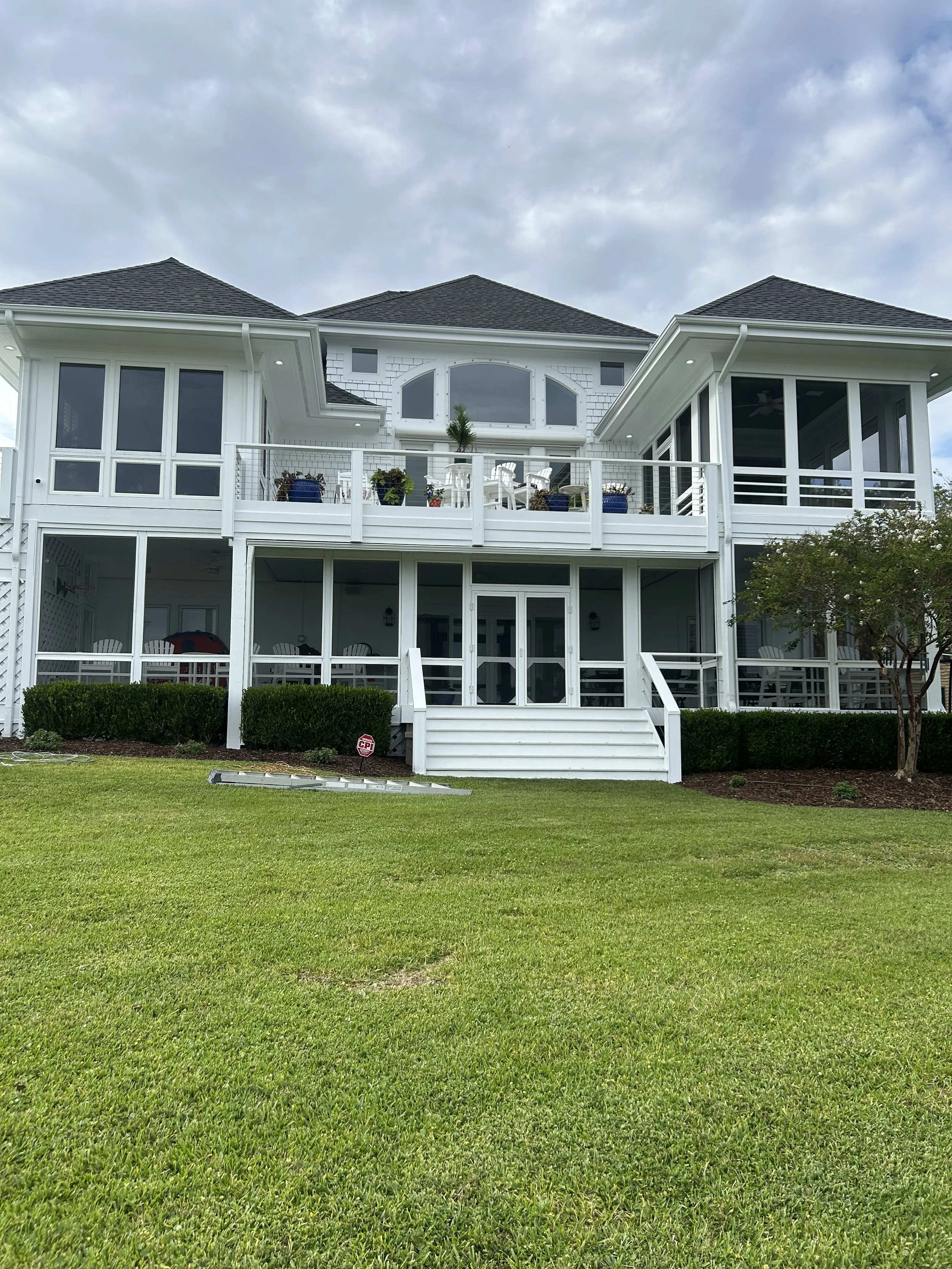 Large white multi-story house with screened-in porches, stairs leading to a grassy yard, and a cloudy sky.