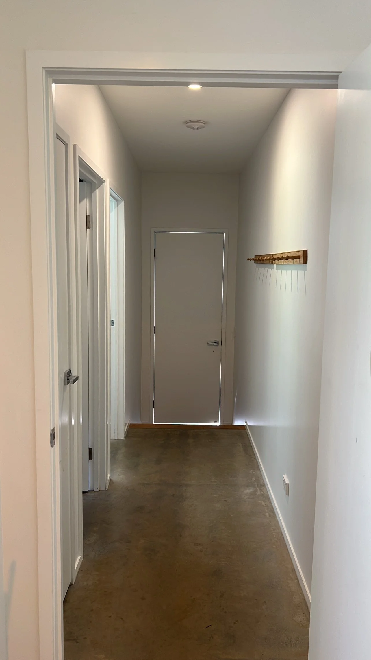 Empty hallway with white walls, a wooden coat rack on the right wall, and a closed door at the end.