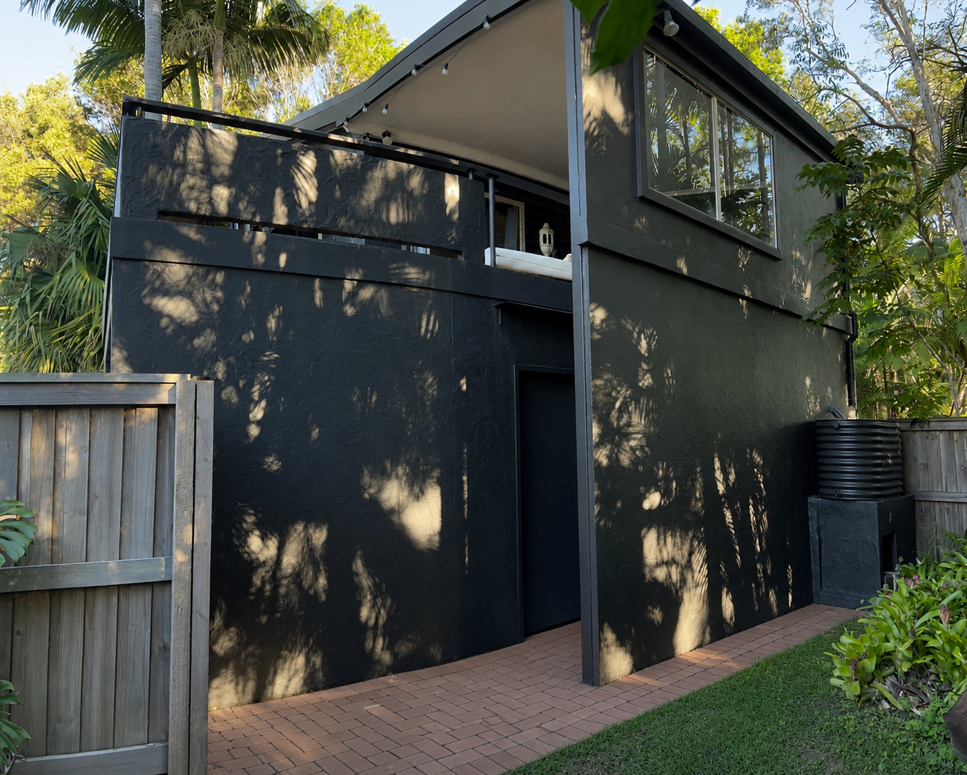 A modern two-story house painted in dark color with large windows, surrounded by trees and greenery, with a brick pathway and wooden fence in the yard.