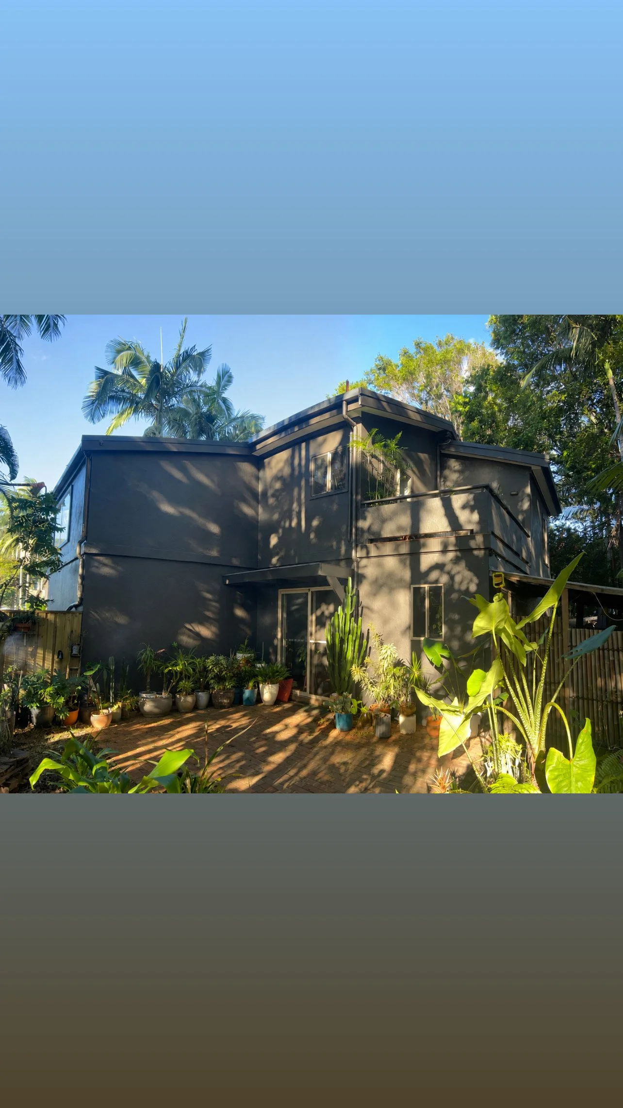 A modern two-story house painted in dark gray, surrounded by lush green trees and plants, with potted plants on a brick patio in the foreground, under a clear blue sky.