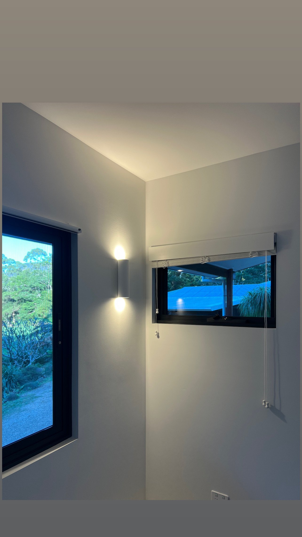 Interior corner of a room with two windows, one on each wall, showing greenery outside. A wall-mounted light is on, casting a warm glow. There is a window blind on one window with a cord hanging down and an electrical outlet at the bottom right.