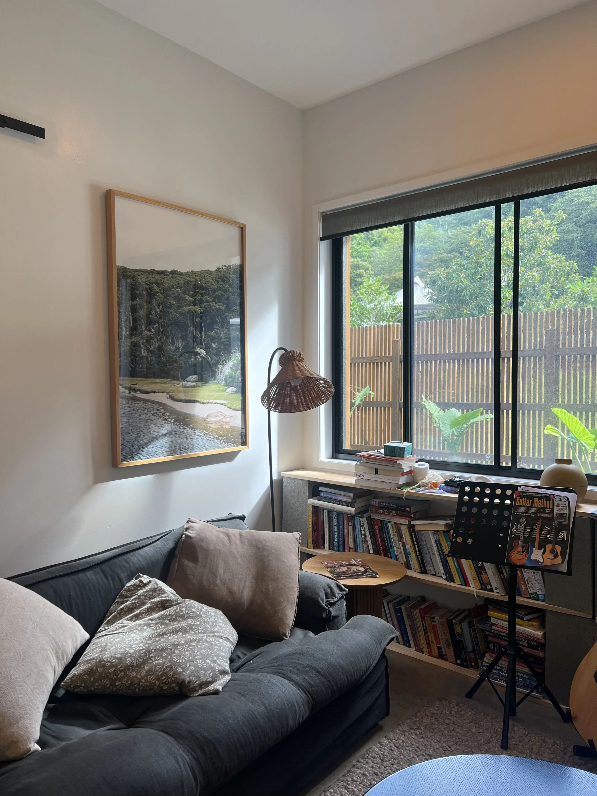 Living room with a dark blue couch, beige pillows, a wooden side table with magazines, a bookshelf filled with books, a lamp with a wicker shade, large window with black frames and a view of a wooden fence and green trees outside, a framed watercolor