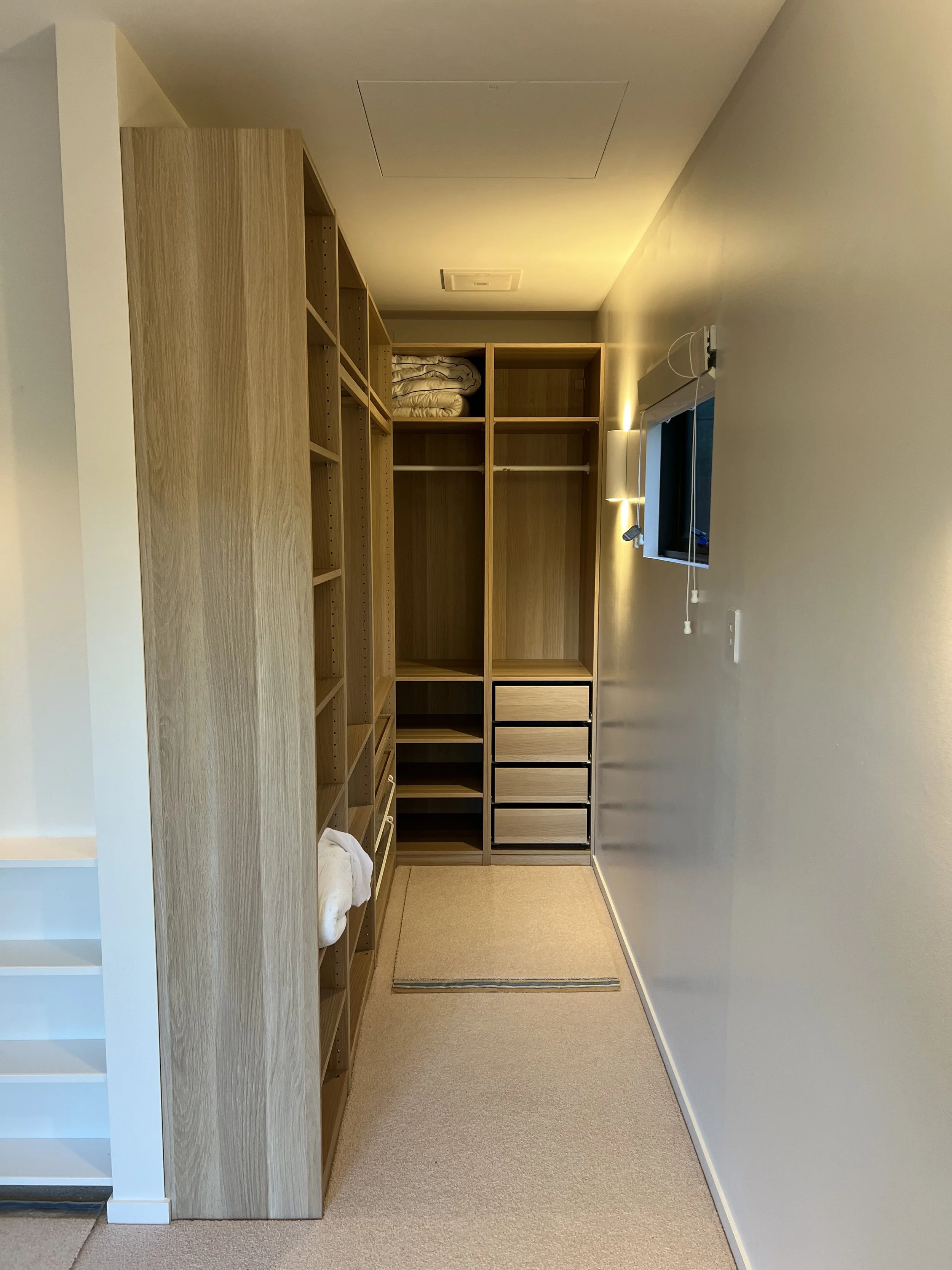 A small walk-in closet with wooden shelving and drawers on the left. The closet has a beige carpet and a window with blinds on the right wall. The ceiling has a light fixture, and the walls are painted in neutral tones.