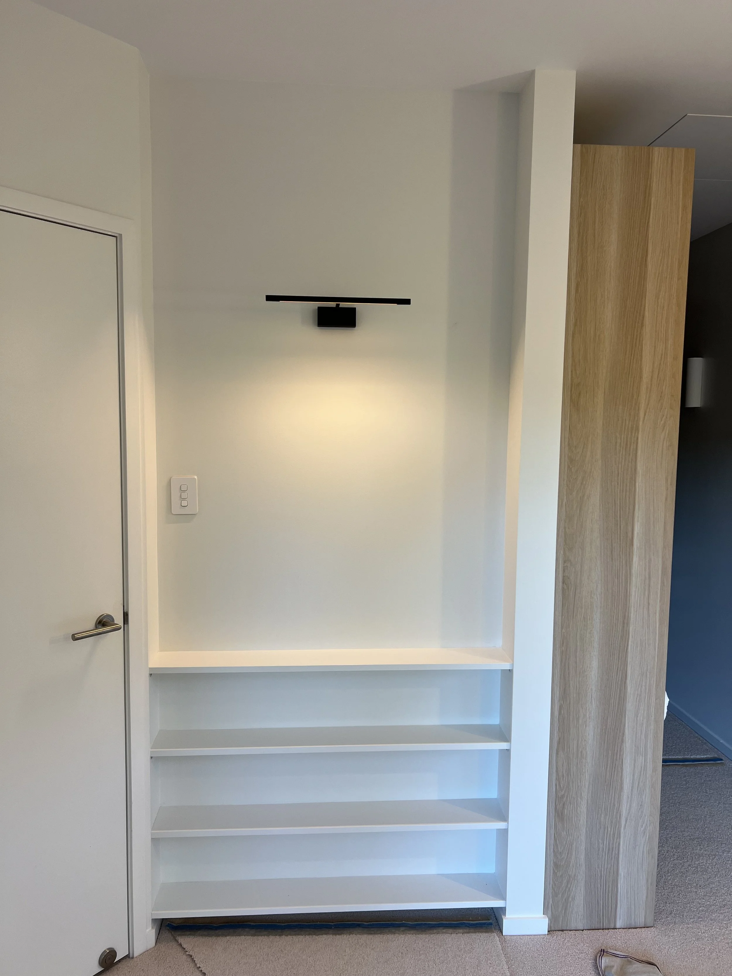 Empty white bookshelf with three shelves next to a wall and a wooden room divider, with a closet door and light switch visible.
