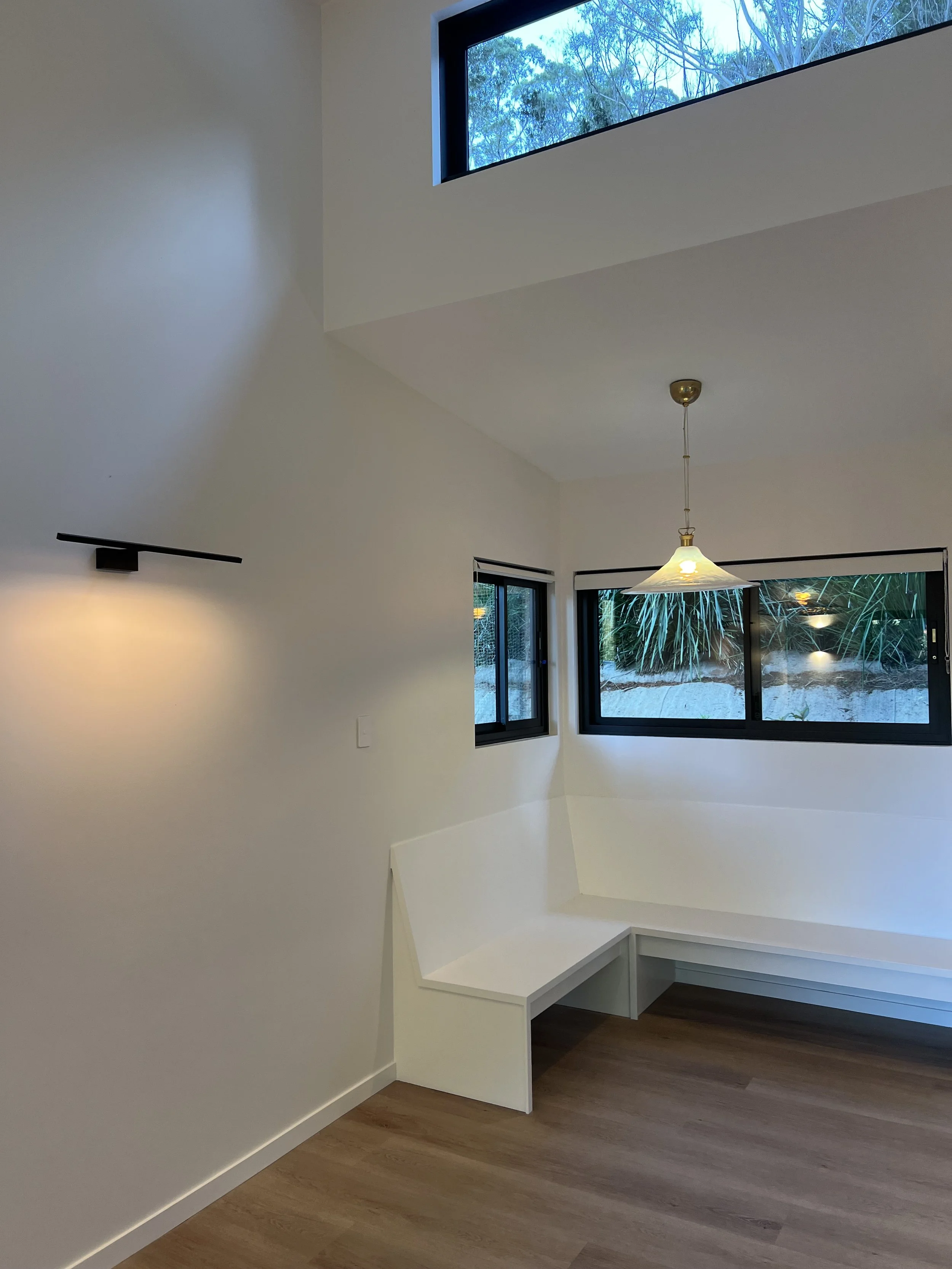 Interior of a modern room with wooden flooring, white walls, a built-in white bench, multiple windows showing outdoor greenery, a hanging light fixture, and a high window near the ceiling.