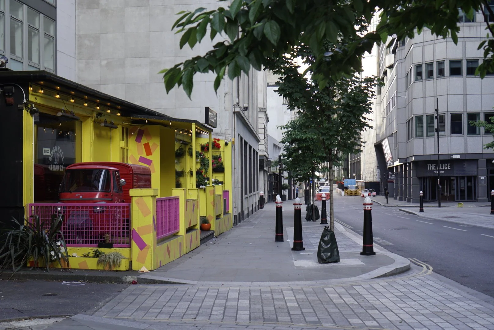 City street scene with a colorful yellow and pink storefront on the left, a red van parked inside, trees lining the sidewalk, and modern high-rise buildings in the background.