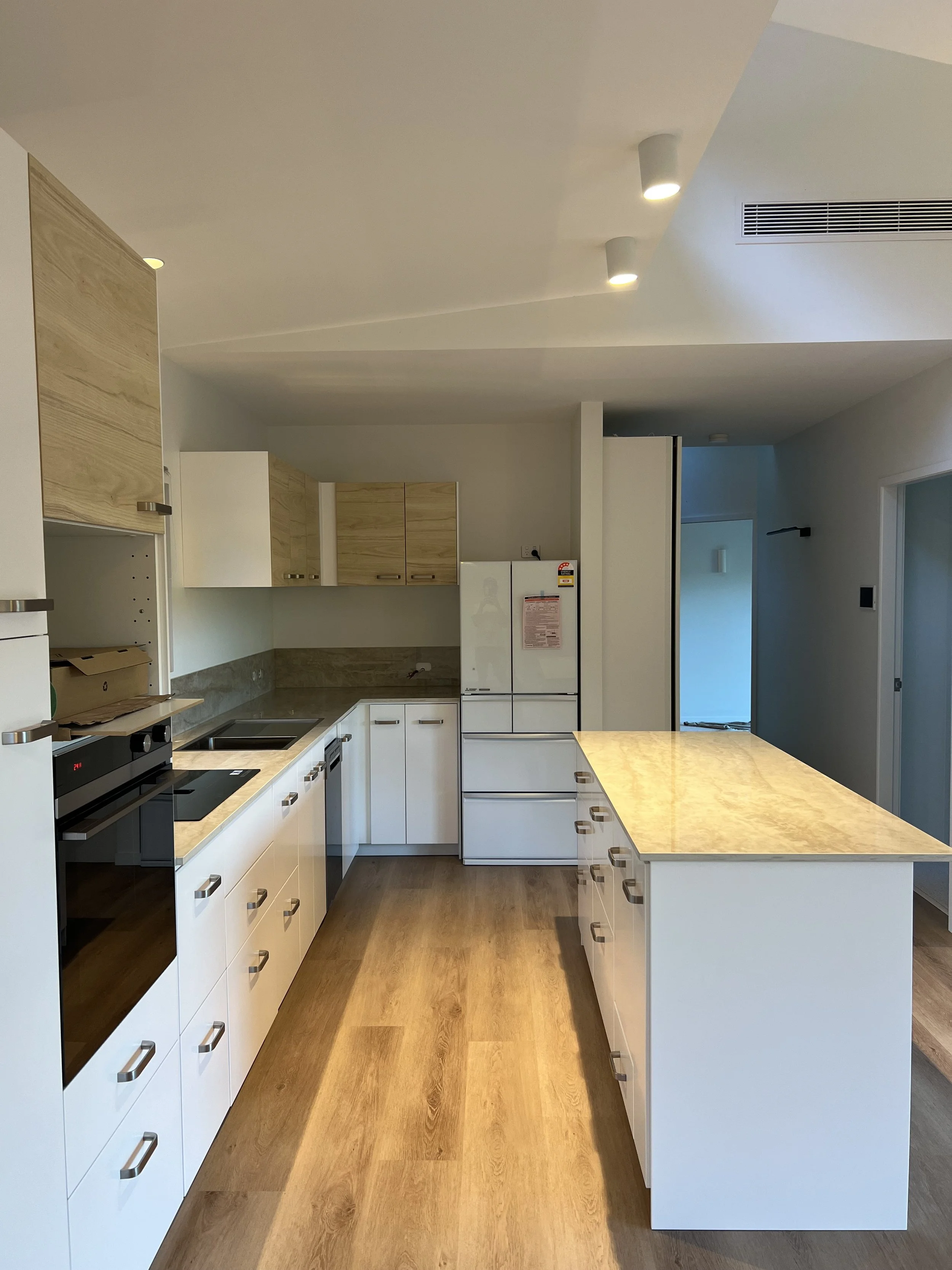 Modern kitchen with white cabinets, wooden upper cabinets, black oven, refrigerator, beige marble countertops, and wooden flooring.
