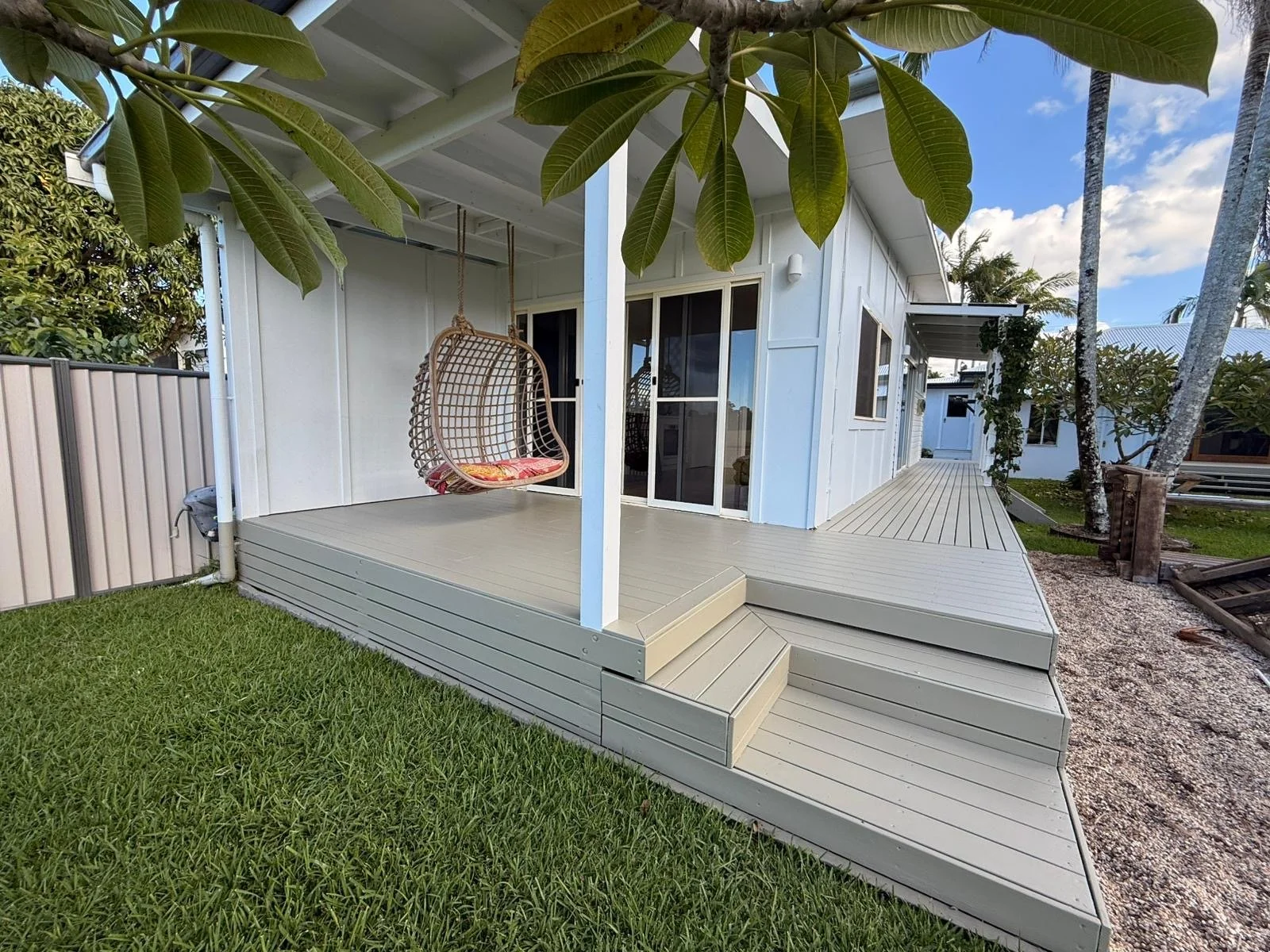Backyard with a wooden deck featuring a hanging wicker swing with cushion, surrounded by green grass, palm trees, and a white house with sliding glass doors and windows.