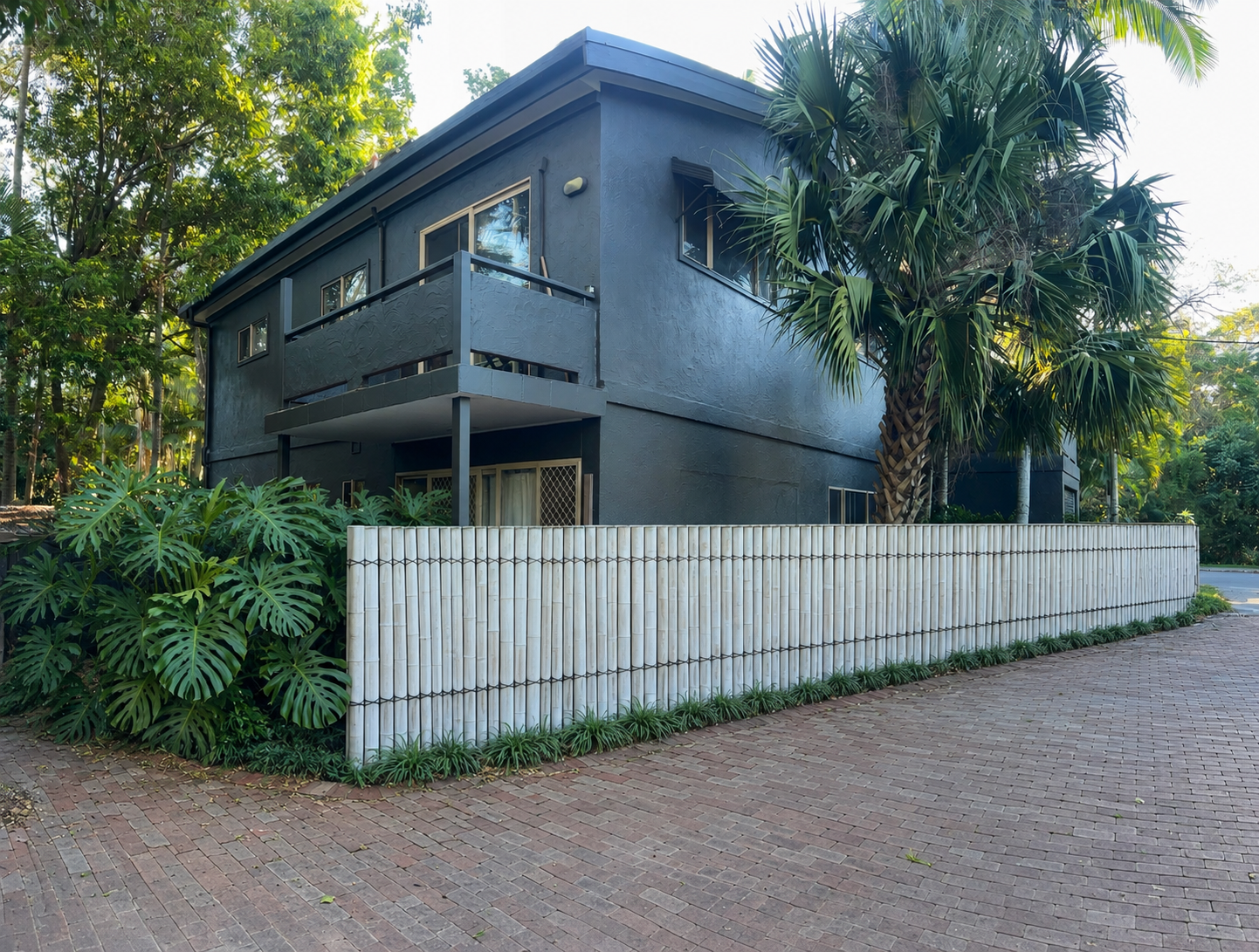 A dark blue two-story house with a balcony, surrounded by lush green tropical plants, including a large palm tree, and enclosed with a white fence, situated on a brick-paved street in a suburban neighborhood.