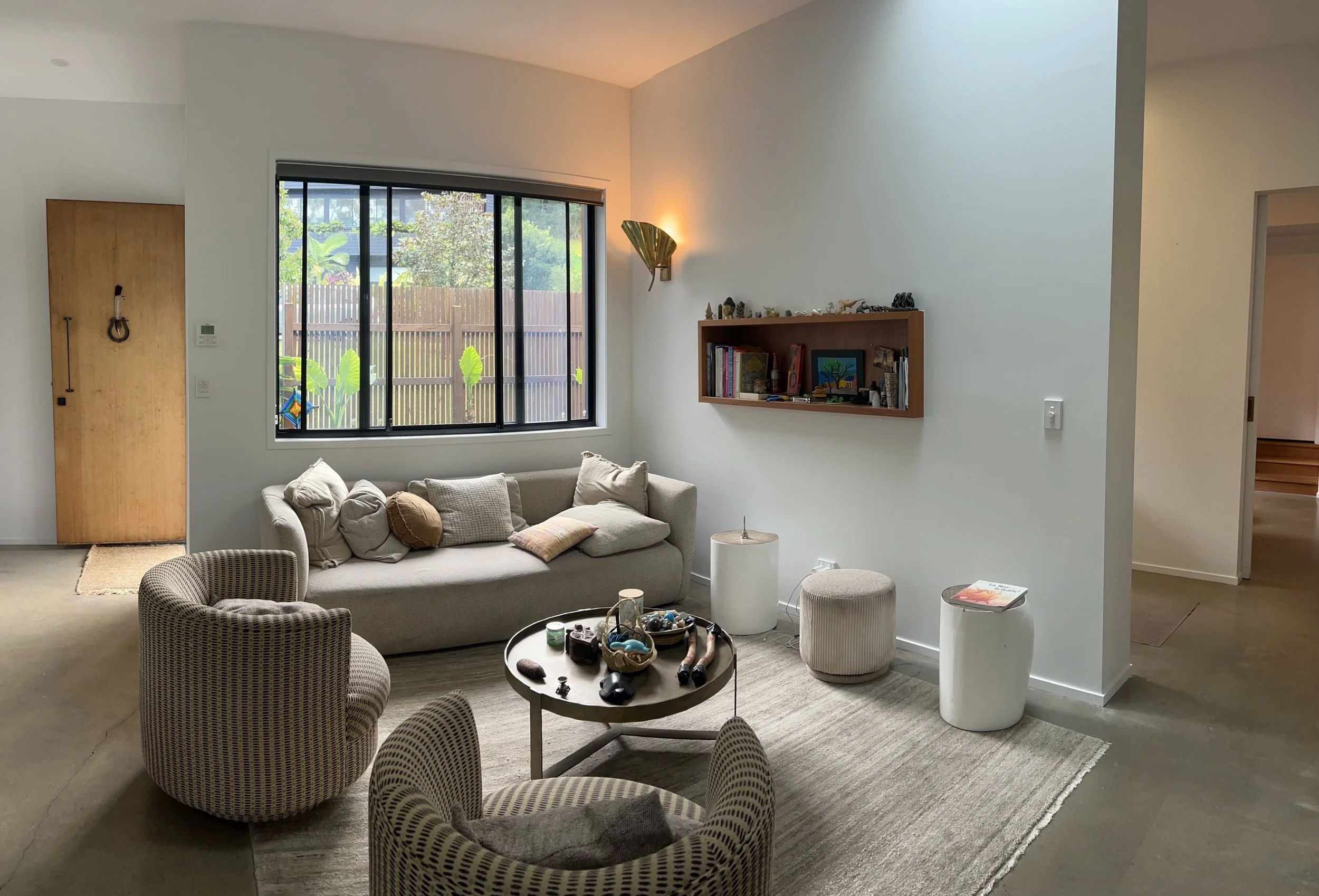 Living room with a large window, sofa with pillows, a wooden shelf with books and decorations, a round table, and a woven armchair.