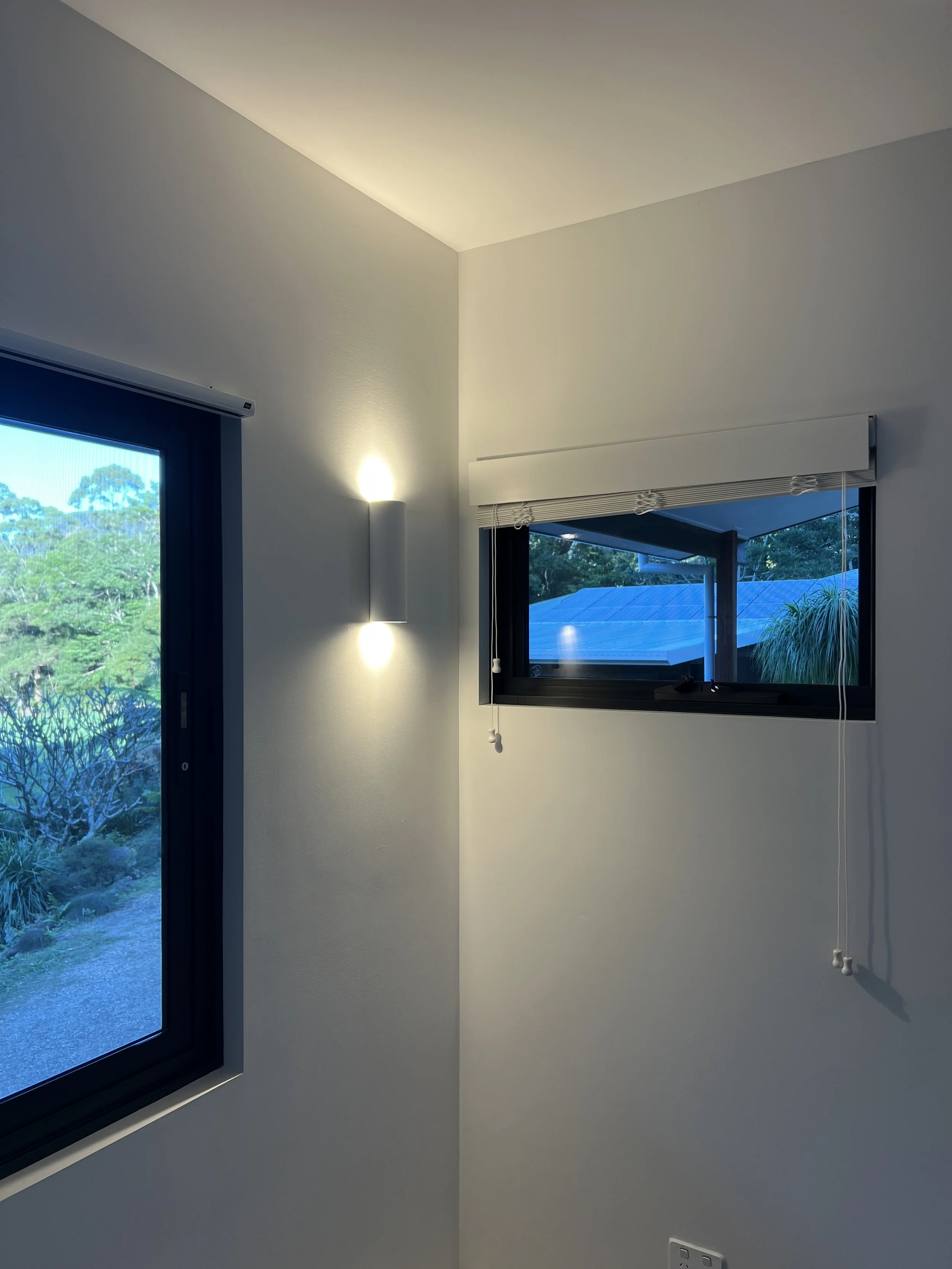 Interior view of a room with two windows, one on each wall, showing trees and a blue sky outside. The wall near the windows has a modern wall sconce with a light. There is a white window blind on the right window with a pull cord hanging down and a w
