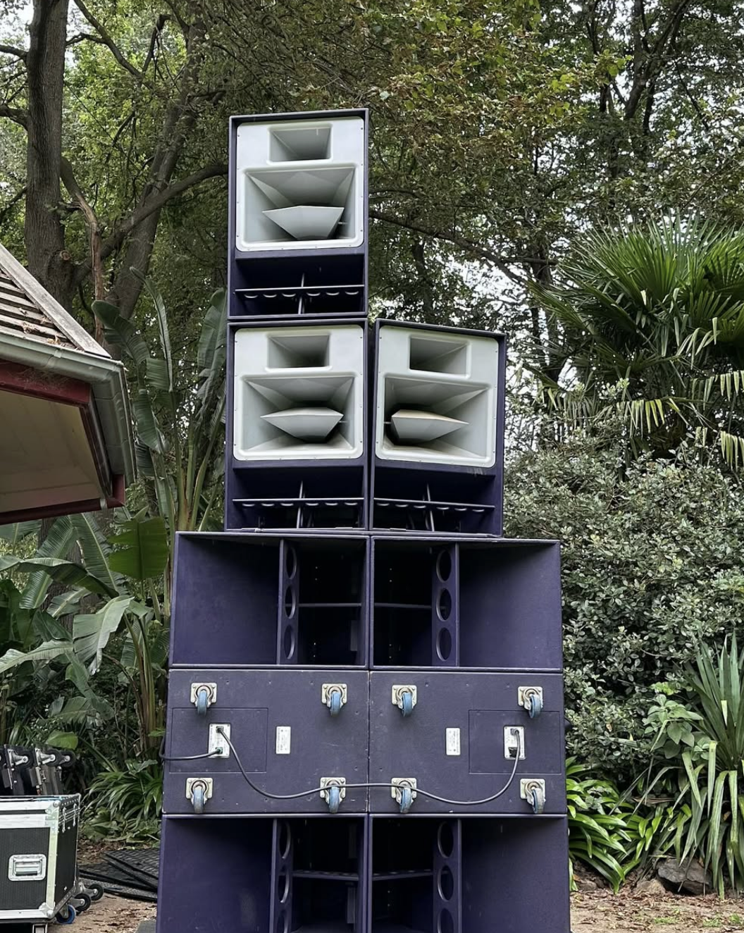 Large outdoor speaker system stacked in a park setting with trees and foliage.