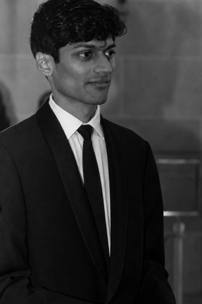 A young man in formal attire, including a black suit, white shirt, and black tie, standing indoors with a neutral expression.