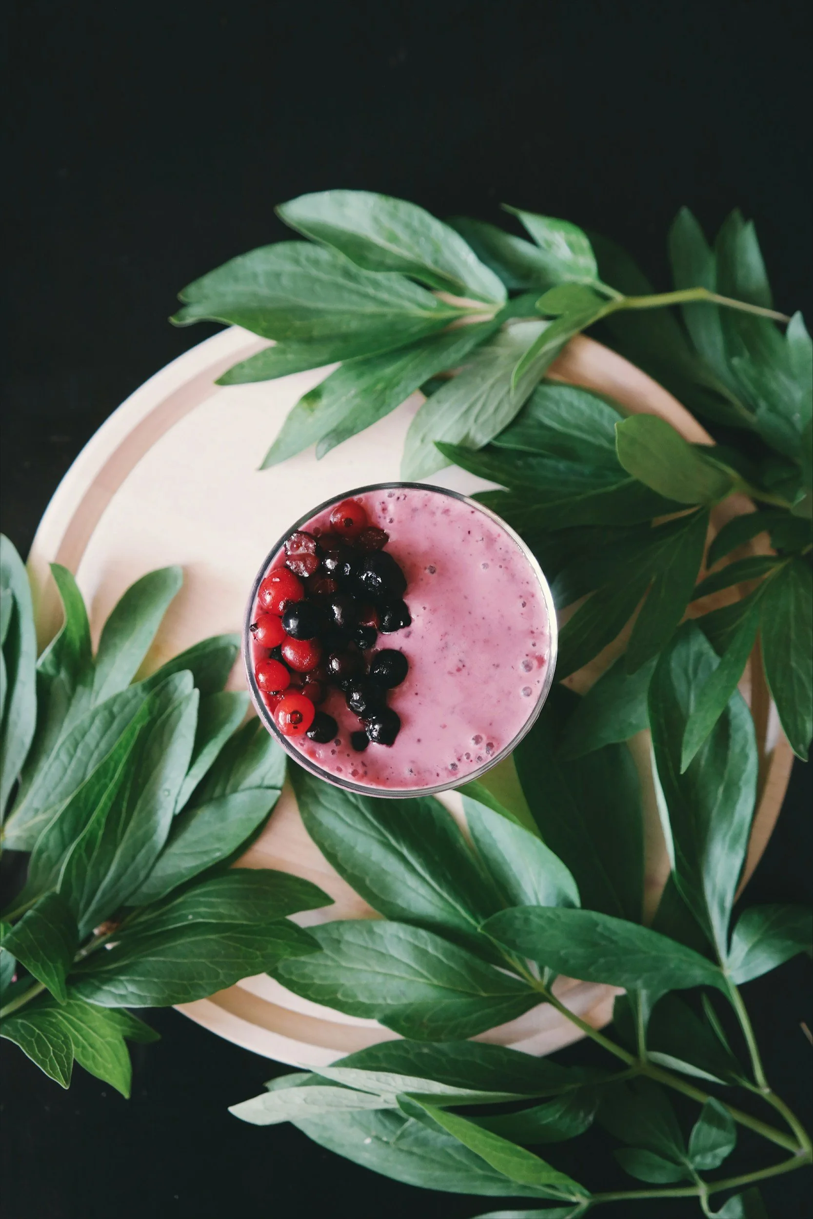 A pink smoothie topped with mixed red and black berries, placed on a round wooden tray surrounded by green leaves.