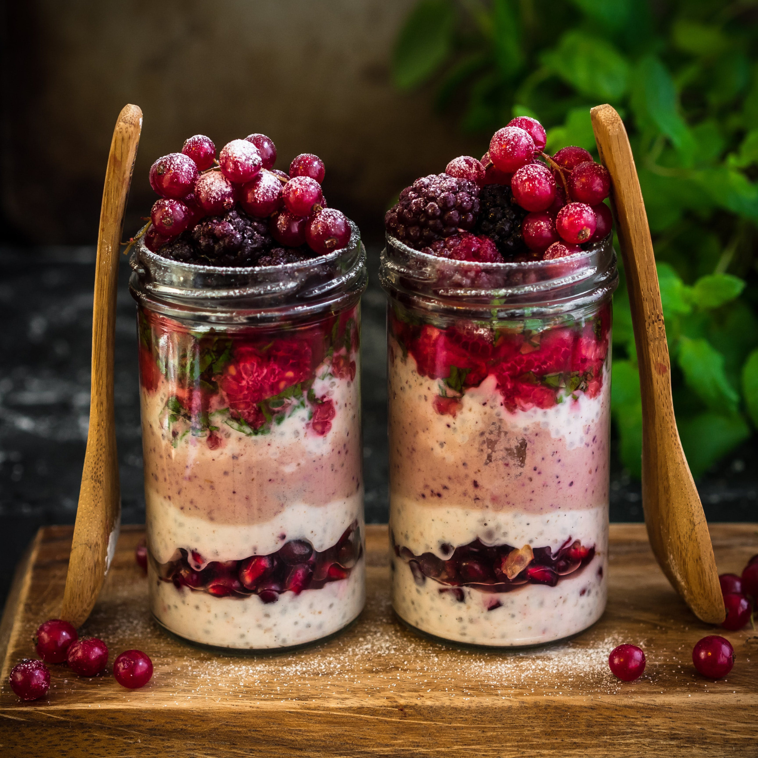 Two glass jars filled with layered berry desserts topped with fresh berries, placed on a wooden tray, with wooden spoons leaning against the jars, and scattered berries around.