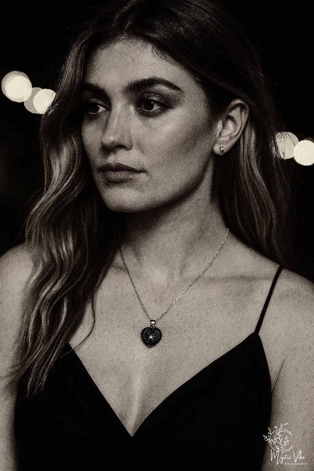 Black and white portrait of a young woman with wavy hair, wearing a necklace with a heart-shaped pendant and a thin-strapped top, against a dark background with bokeh lights.