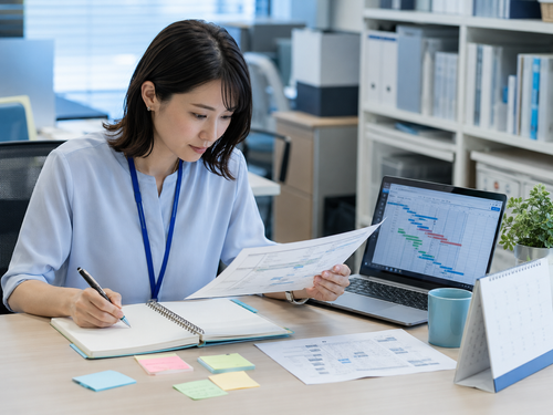 A project coordinator reviewing printed schedules and a project timeline on a laptop.