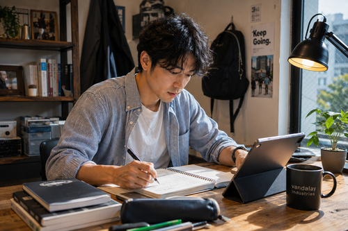Man writing in a notebook beside an open laptop at a study desk
