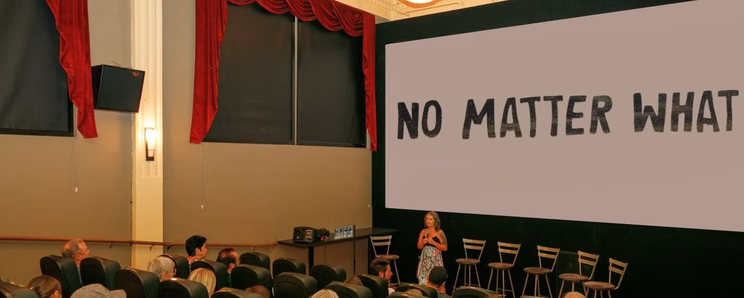 A woman in a dress stands in front of a large screen that says NO MATTER WHAT