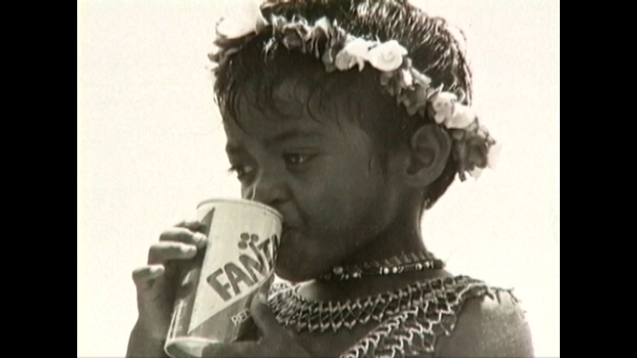 A black and white photo of a young child with flowers in hair drinks from a plastic cup labeled Fanta
