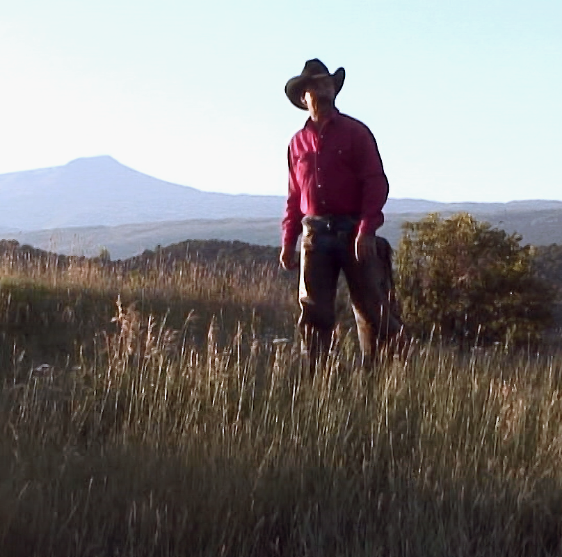 A man in a cowboy hat and button down shirt walks in a field with mountains behind him.