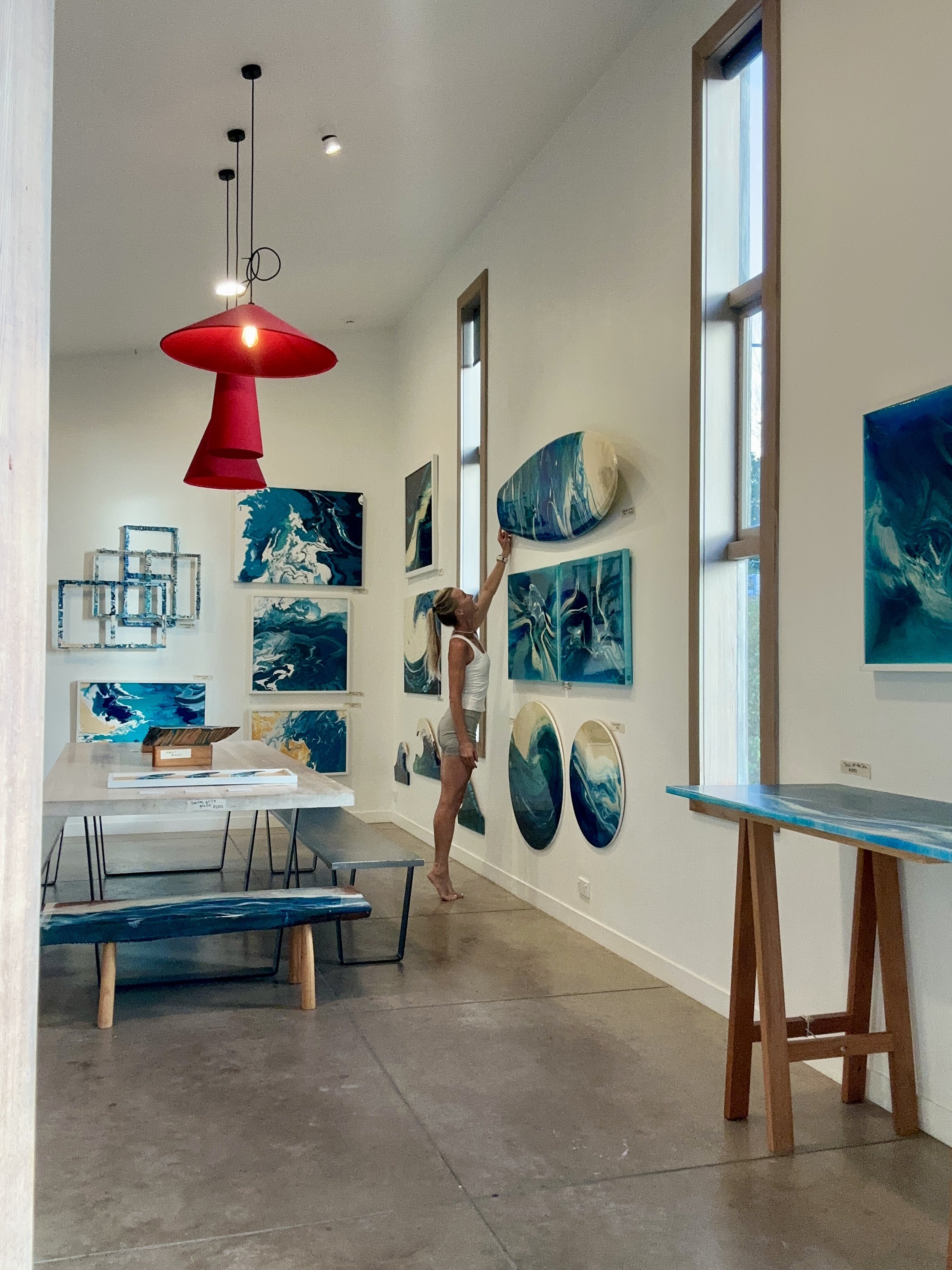 An art gallery with blue and white abstract paintings on white walls, a woman stretching upward, and natural light coming through tall windows.