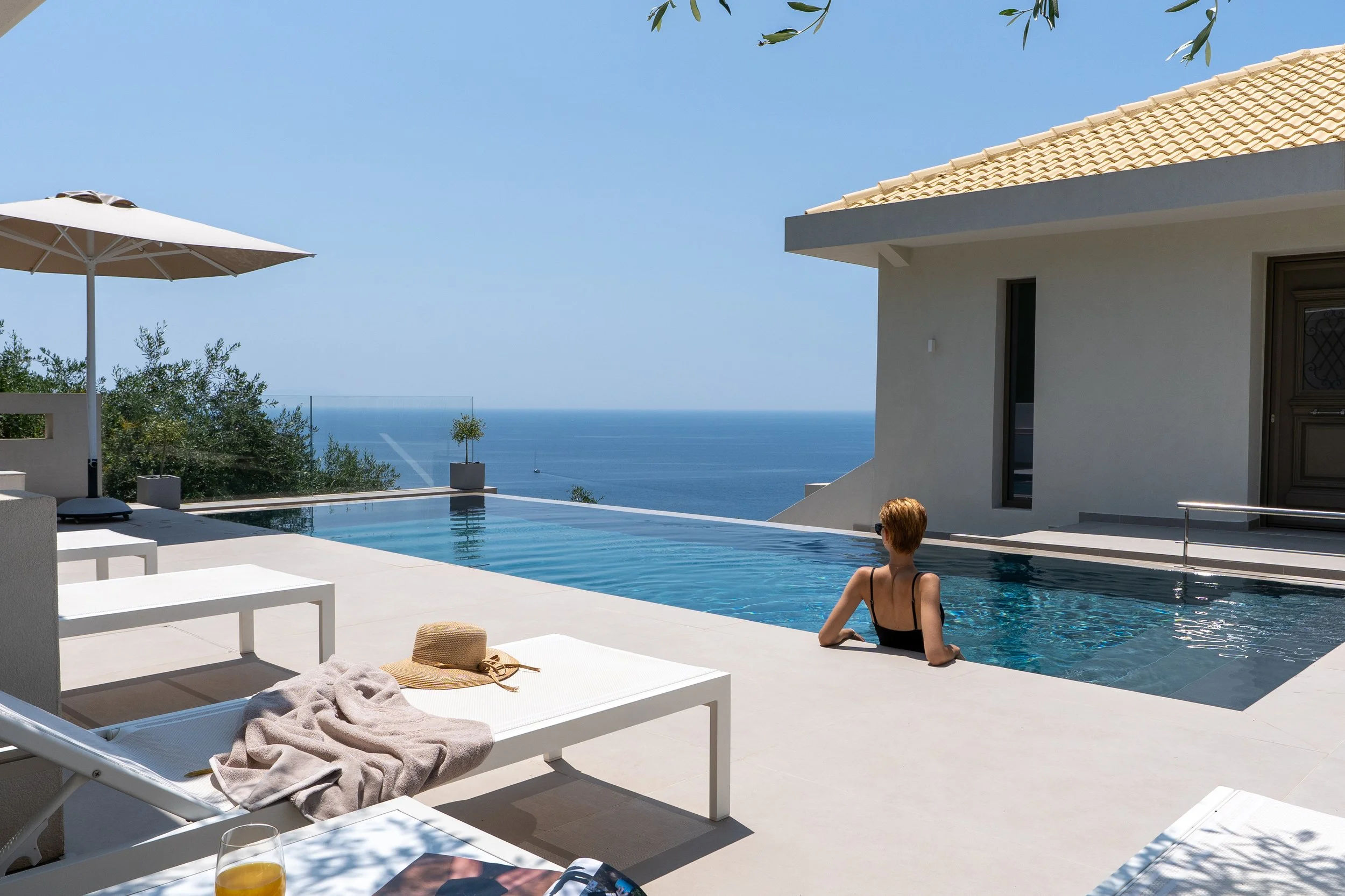A woman relaxing in a swimming pool at a modern house with an ocean view, partly shaded by a large umbrella, with a straw hat, towel, and drink on a poolside lounge chair.