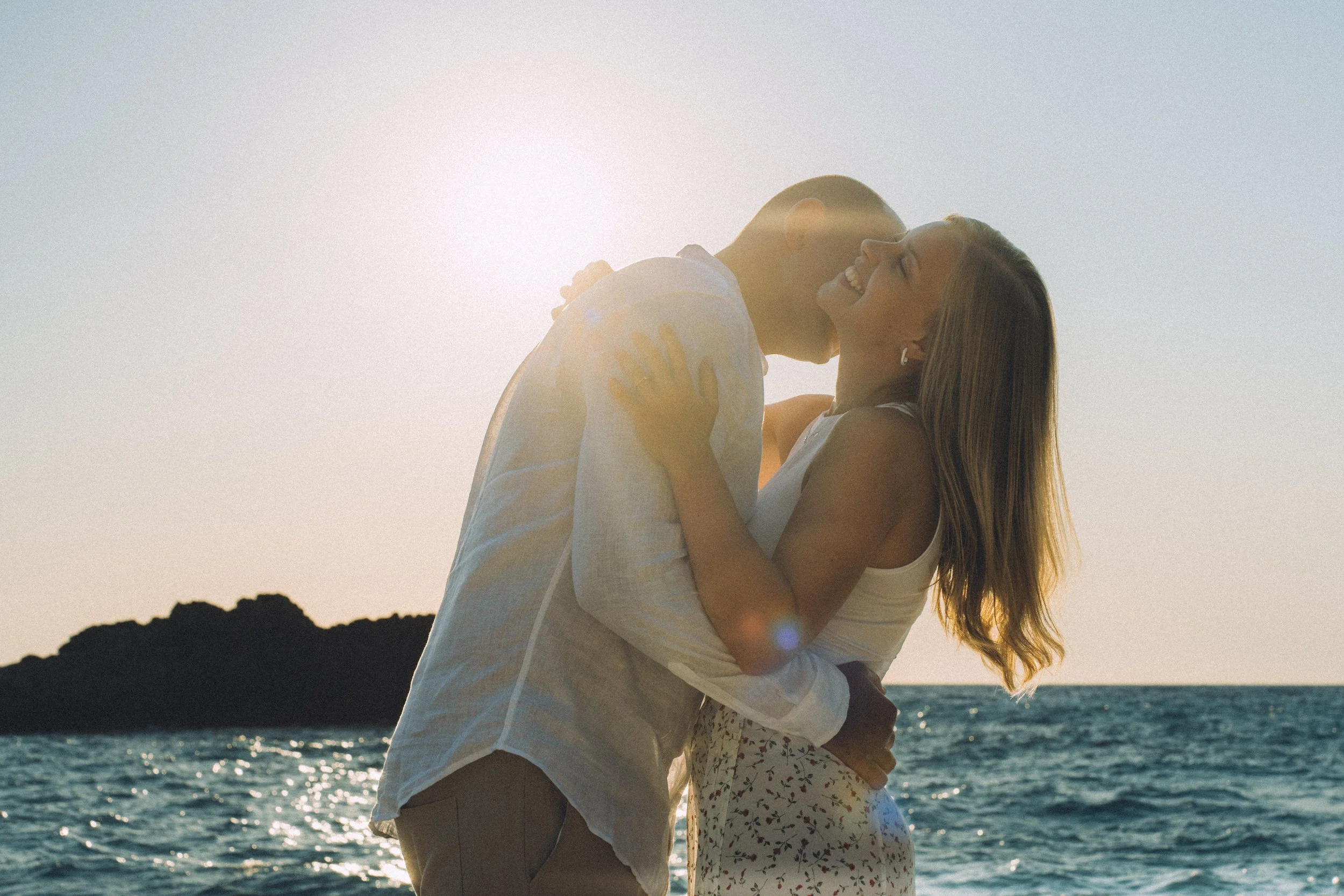 A happy couple embracing and kissing near the ocean at sunset.