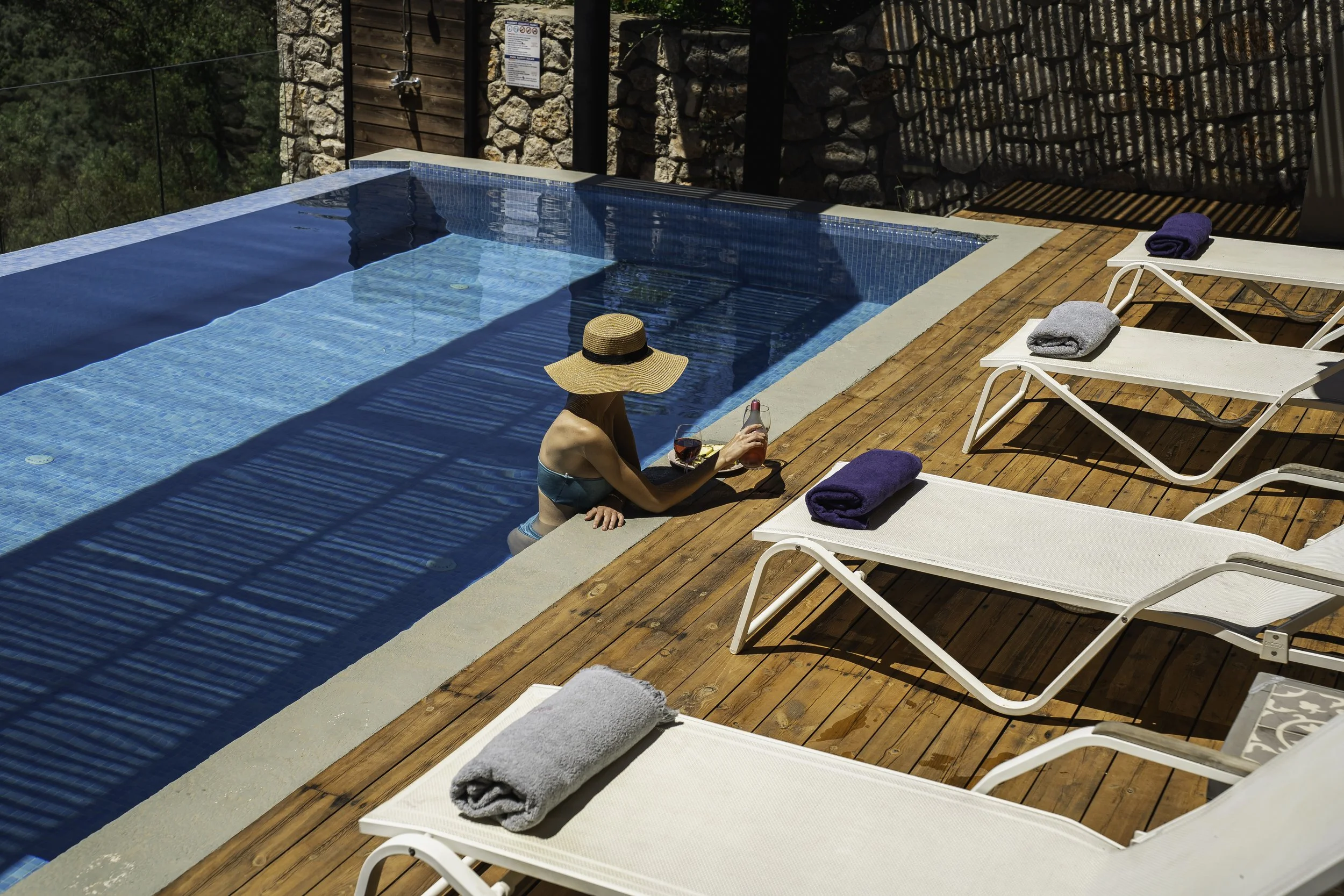 A woman in a bikini and a wide-brimmed hat sits at the edge of a swimming pool, holding a bottle of rosé wine. Surrounding her are poolside lounge chairs with towels, in a sunny outdoor setting.