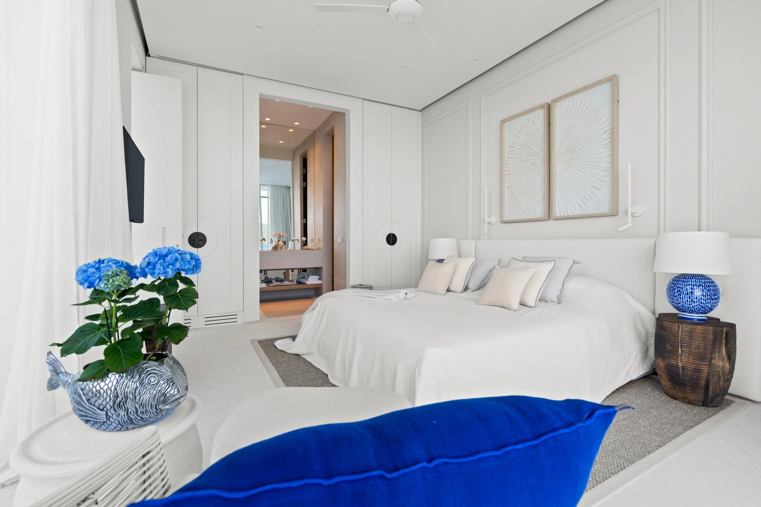 A luxurious hotel bedroom with white walls, a large white bed with multiple pillows, a bedside table with a blue decorative lamp, a flat-screen TV, blue hydrangea flowers in a fish-shaped vase, and a view into the bathroom with a sink and mirror.