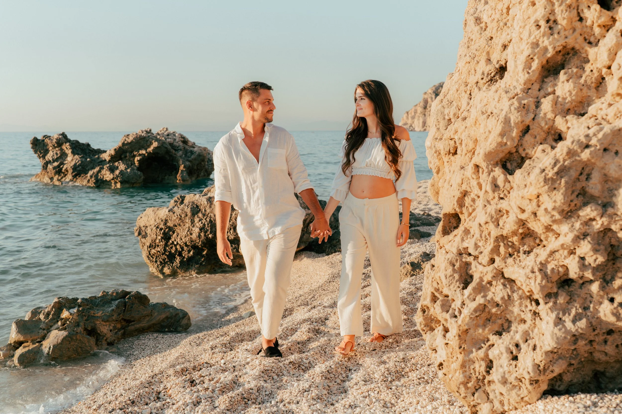 A couple holding hands walking on a rocky beach with large rocks and cliffs, near the ocean under a clear sky.