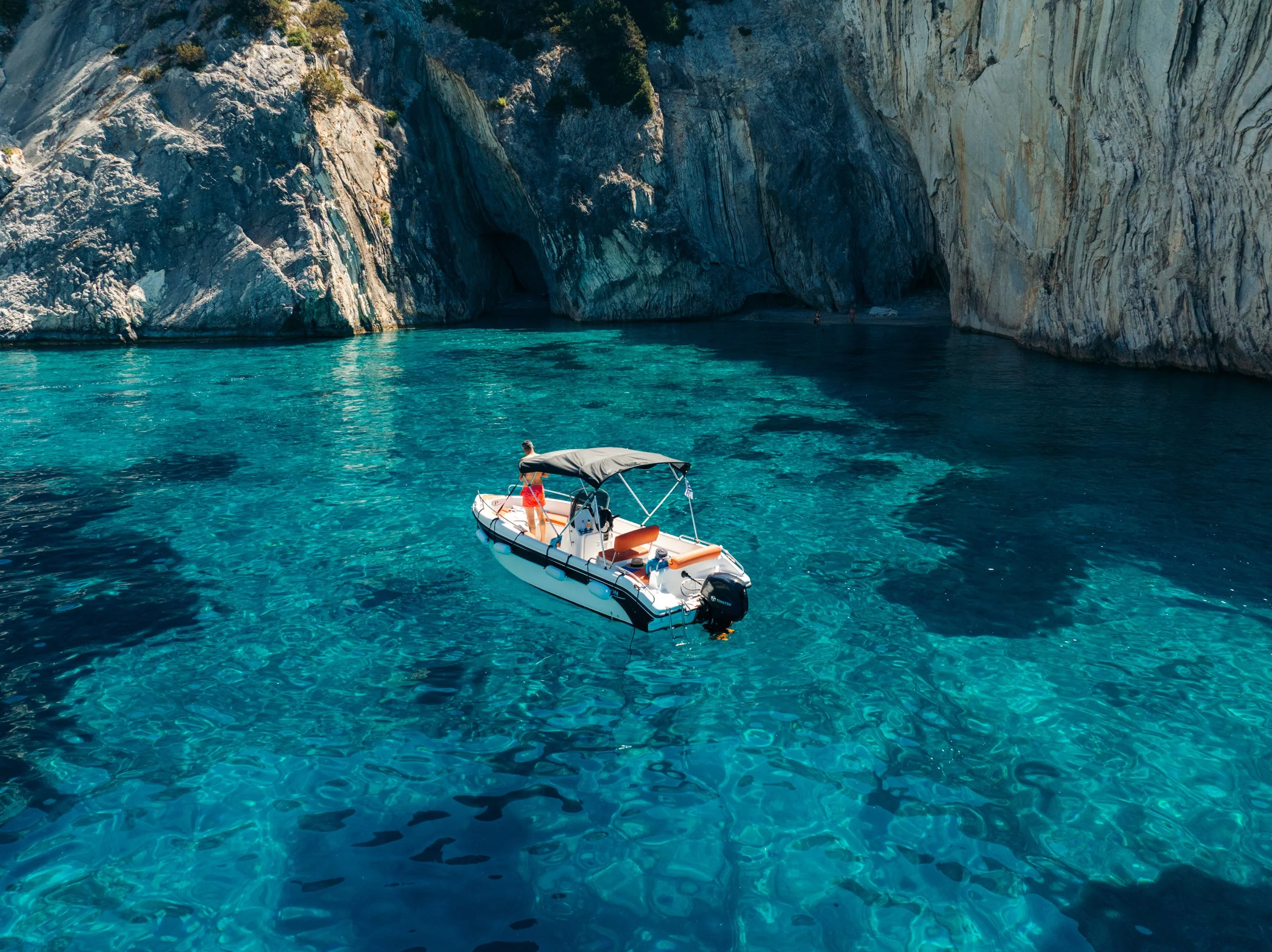 A boat with a person standing on it in clear, turquoise water near rocky cliffs.