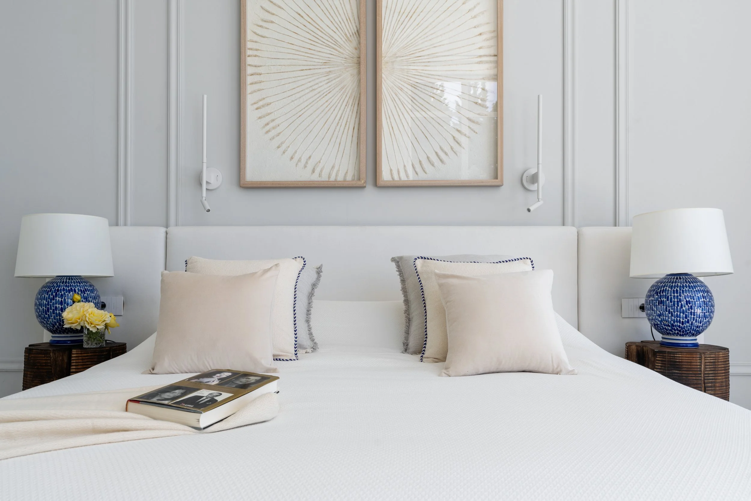 A neatly made bed with a white headboard, four cream pillows with navy striped borders, two blue and white table lamps on wooden nightstands, and modern wall art above the bed in a light-colored room.