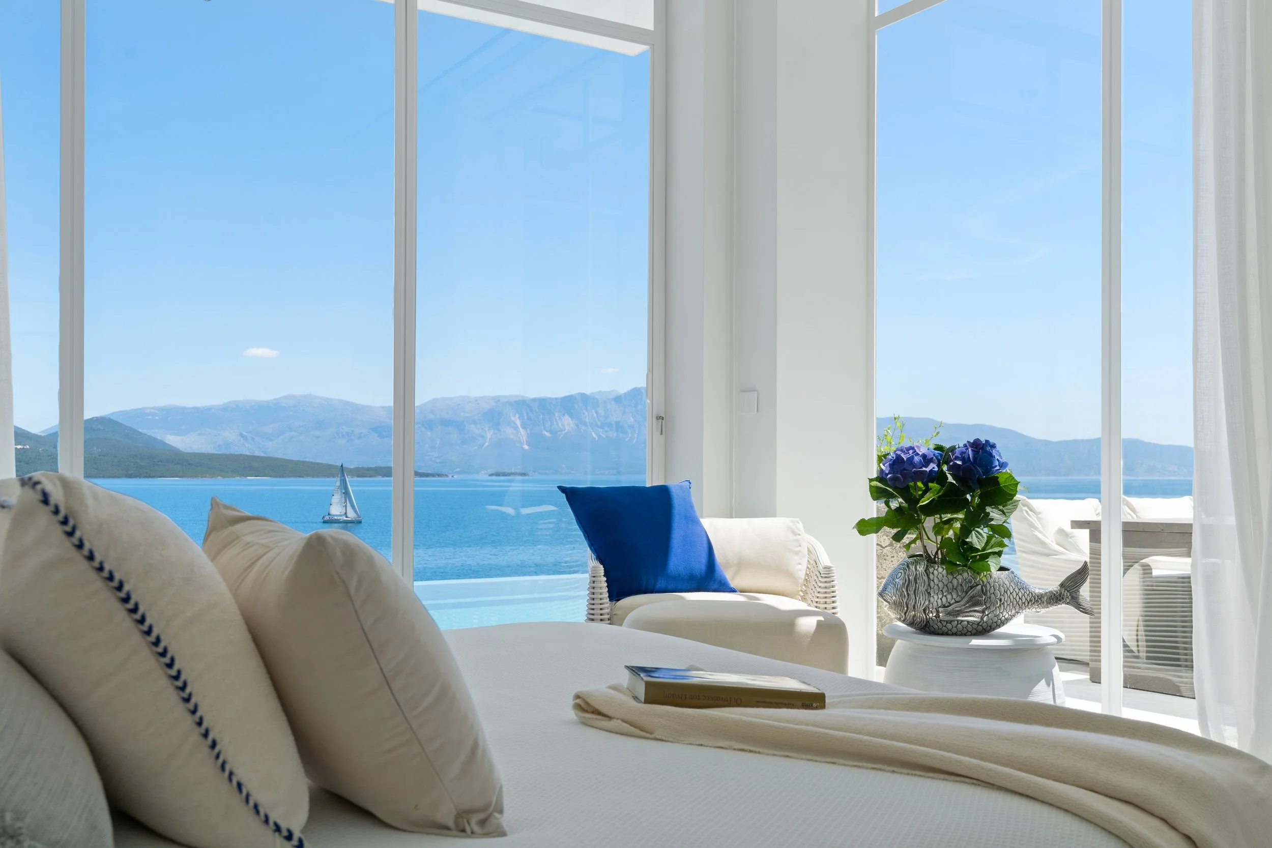 Living room with large glass windows overlooking a lake and mountains, featuring white furniture, a blue pillow, a book, a flower arrangement in a fish-shaped vase, and sheer curtains.