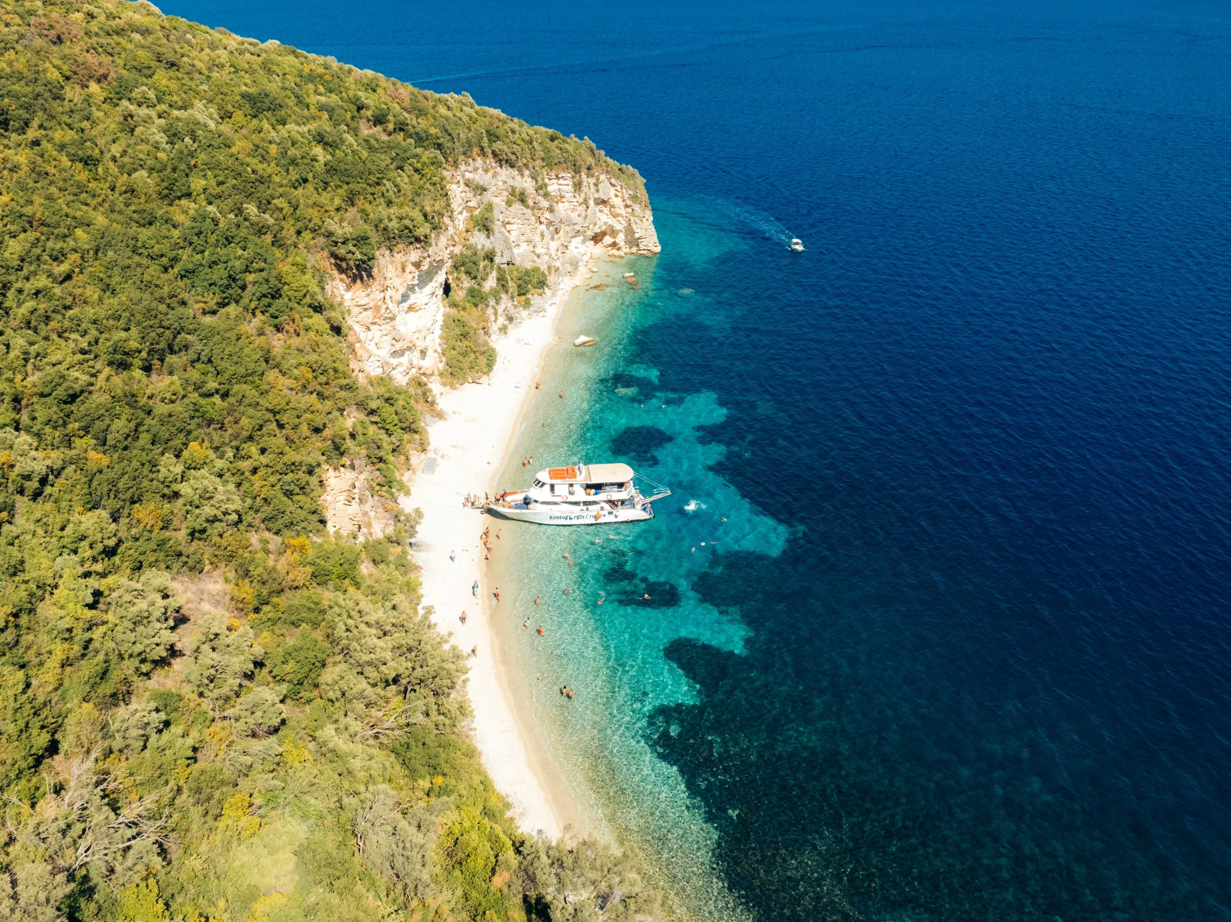 Aerial view of a small beach with a boat anchored near the shore, surrounded by lush green hills and clear blue water.