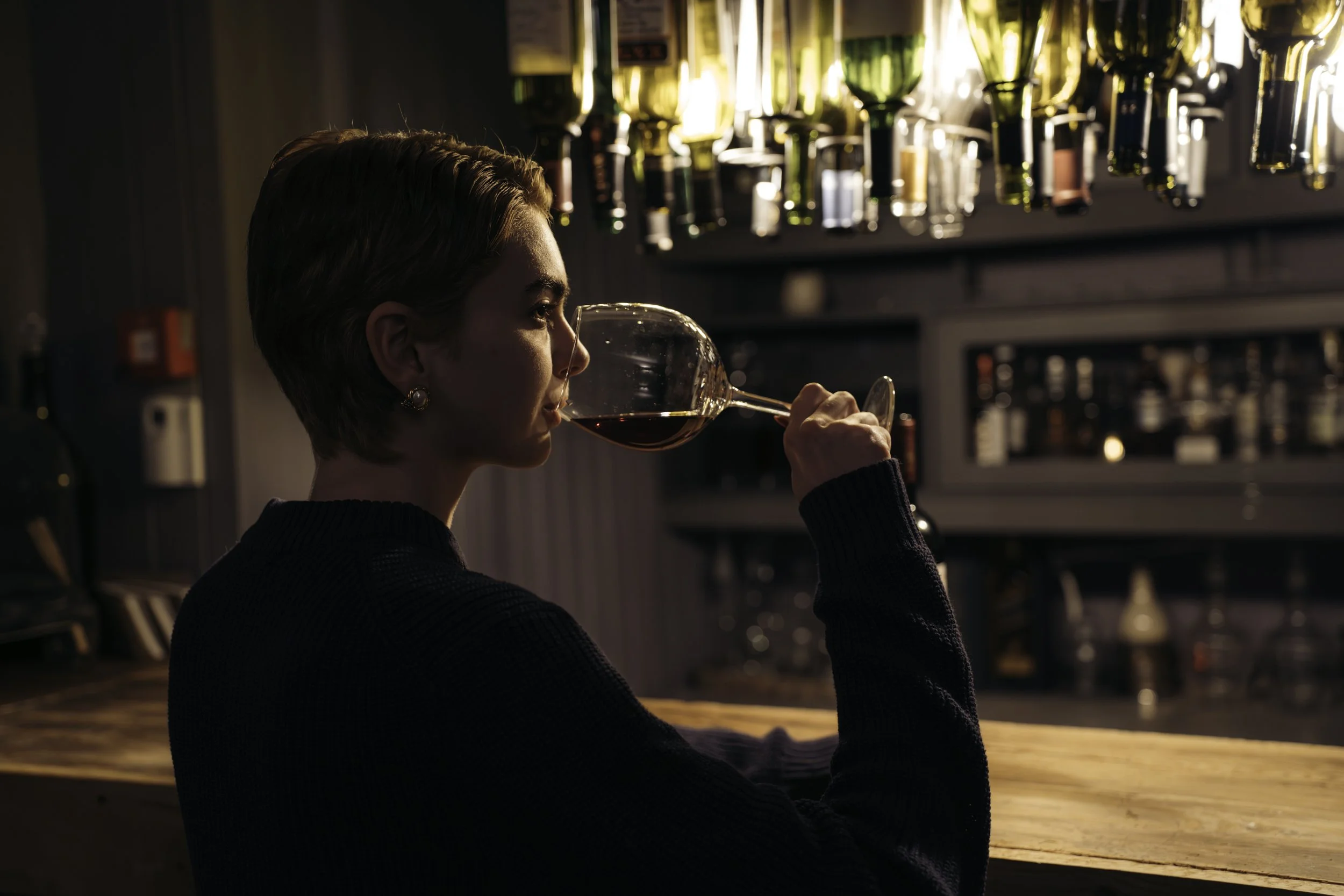 A woman with short hair and earrings, wearing a black sweater, sitting at a bar, holding a glass of red wine near her lips, with a background of bottles on shelves.