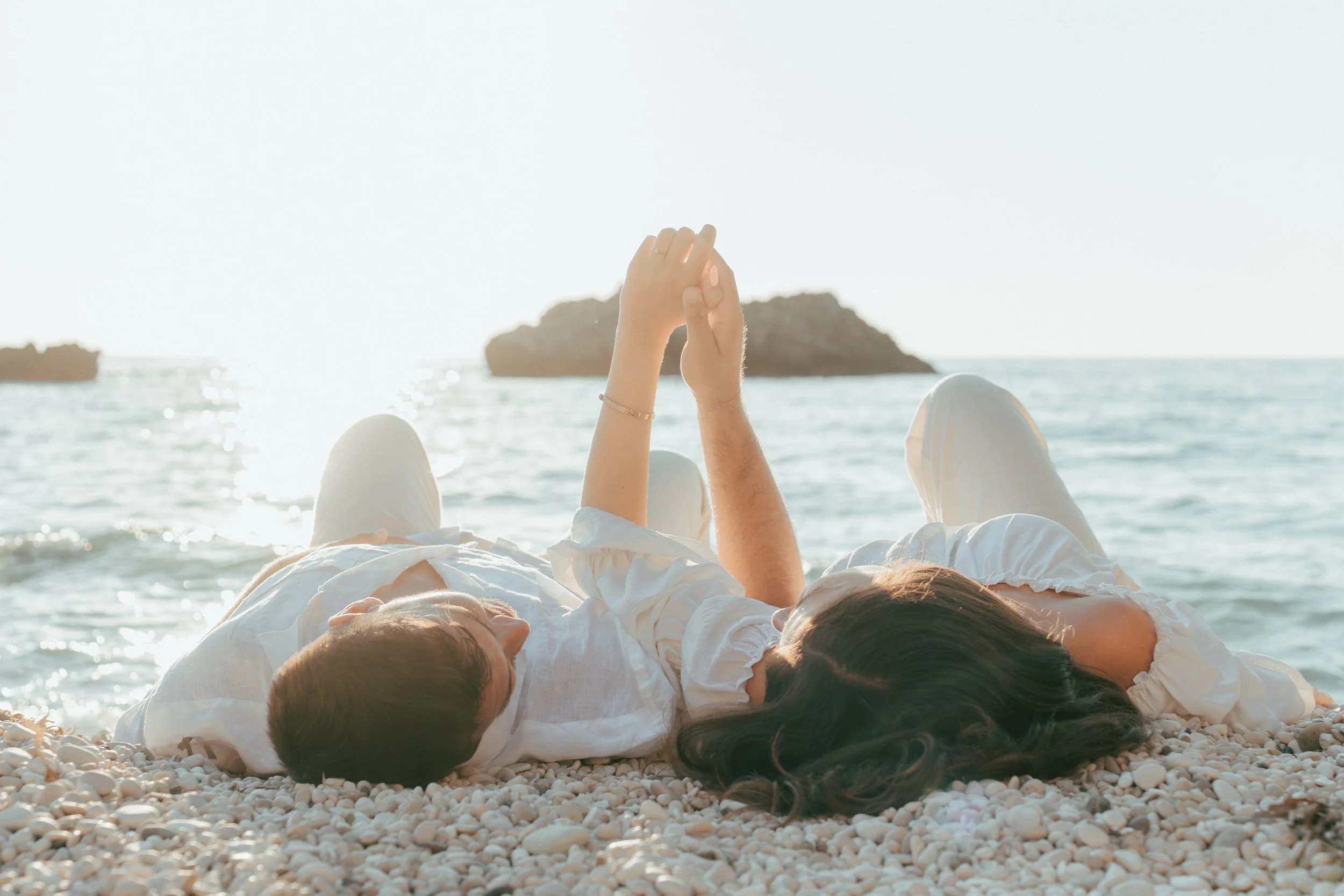 Couple lying on a pebble beach by the sea, holding hands and relaxing at sunset.