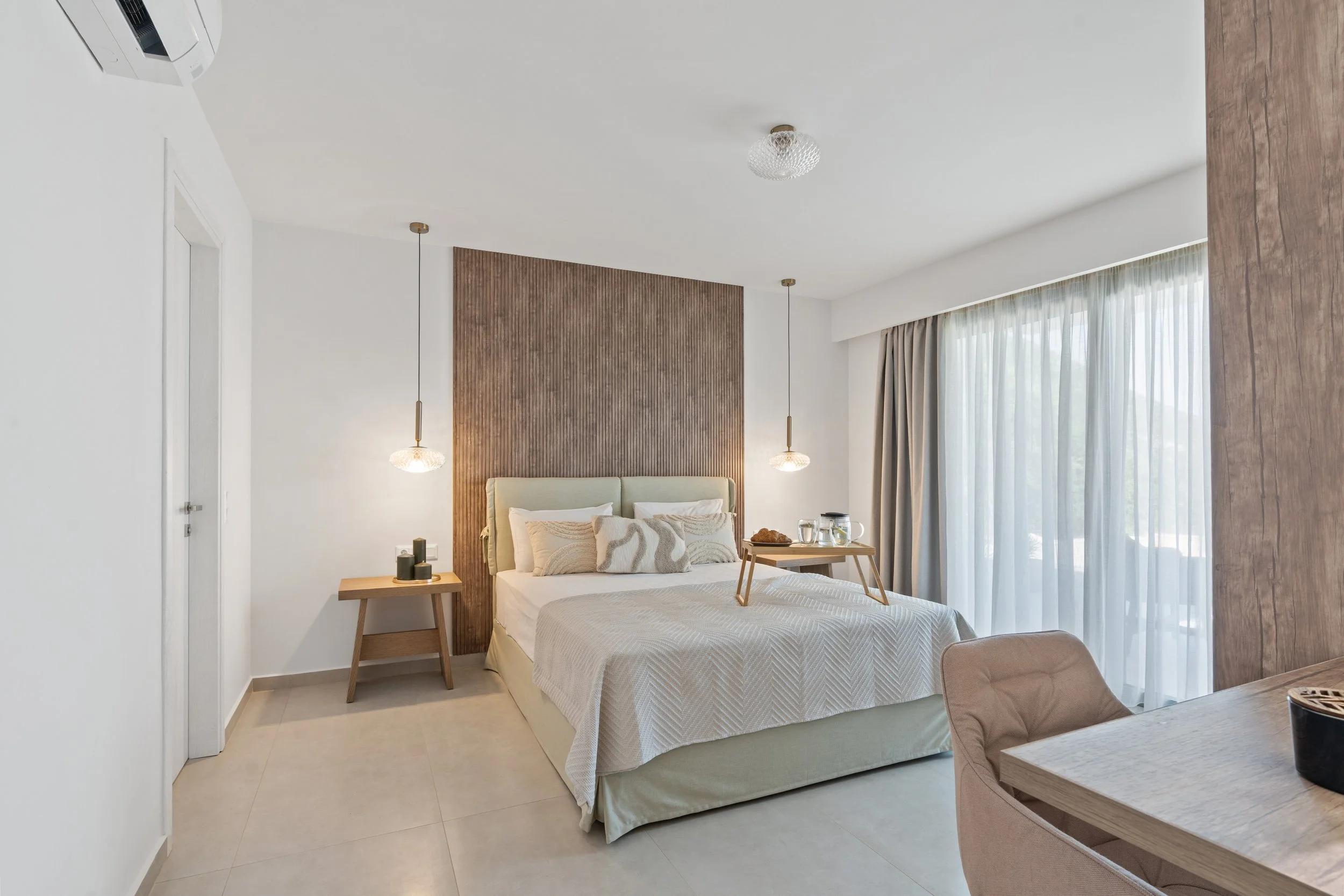 A modern hotel bedroom with a bed, bedside tables, pendant lights, a window with curtains, and a small table with tea-making supplies.