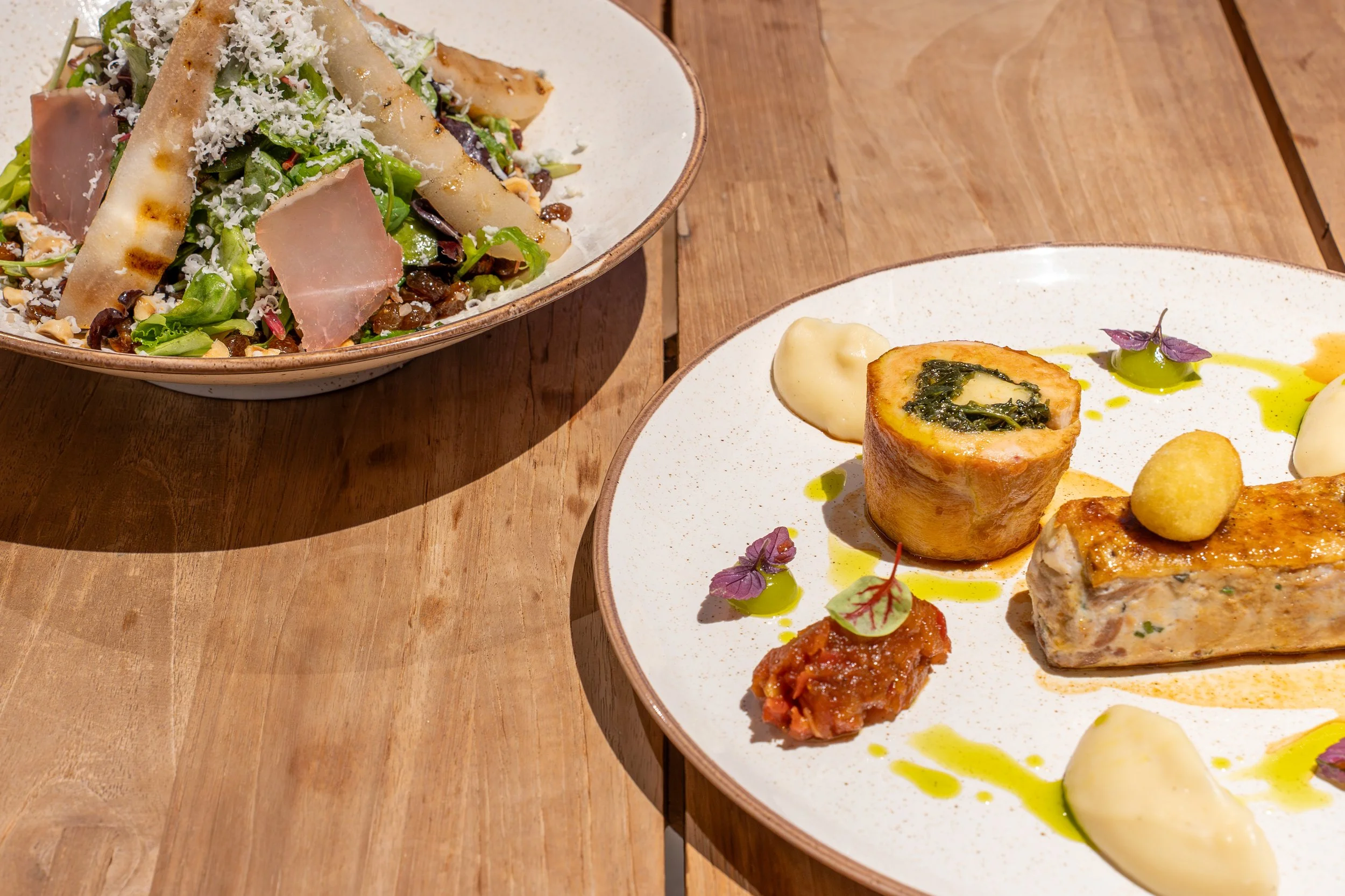 A plate of assorted gourmet food items, including a rolled pastry with green filling, a rectangular piece of meat with white sauce, a small dollop of red sauce garnished with a microgreen, and various drizzled sauces with edible purple microgreens on