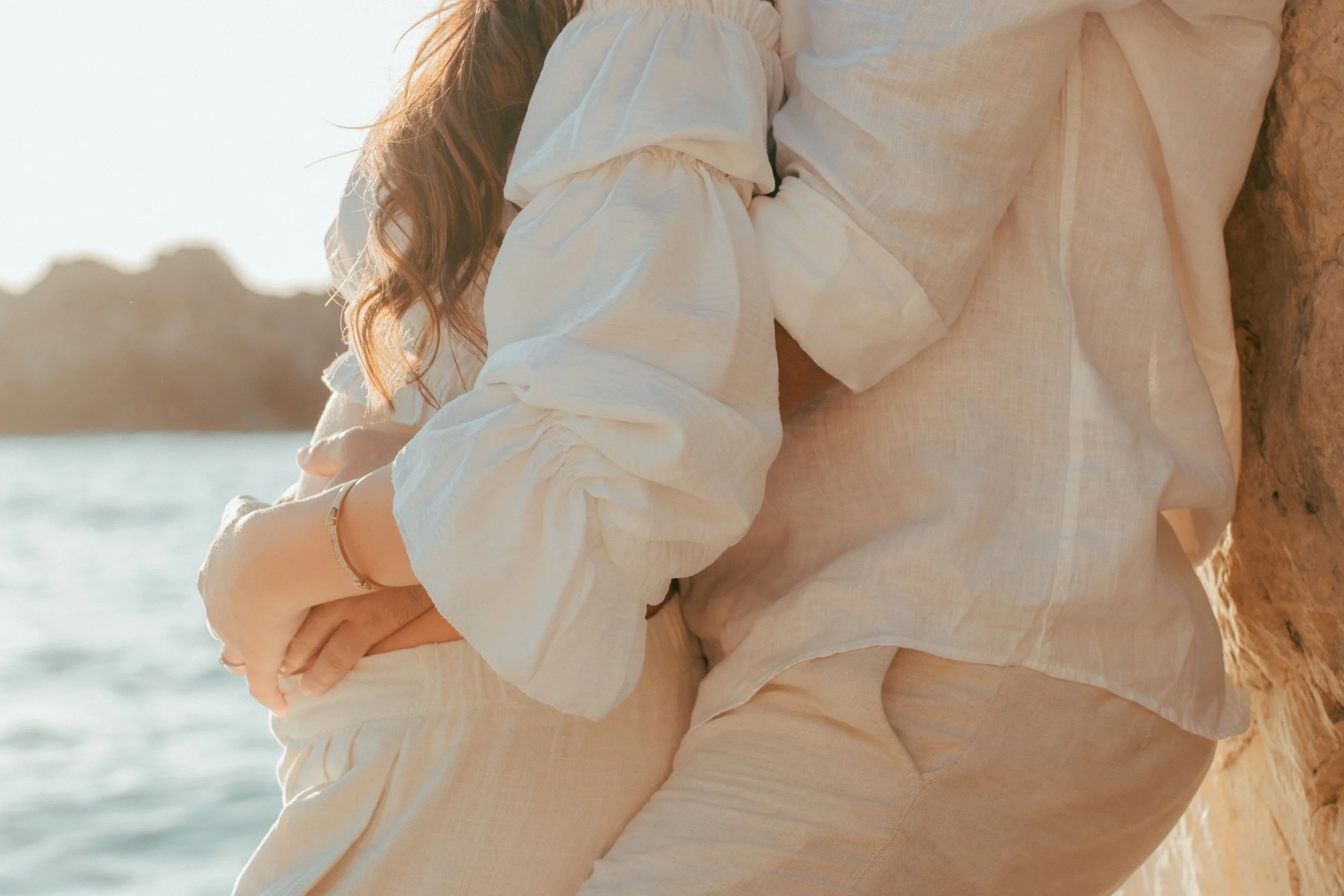 A person in light-colored clothing sitting against a rocky surface by the water during sunset.