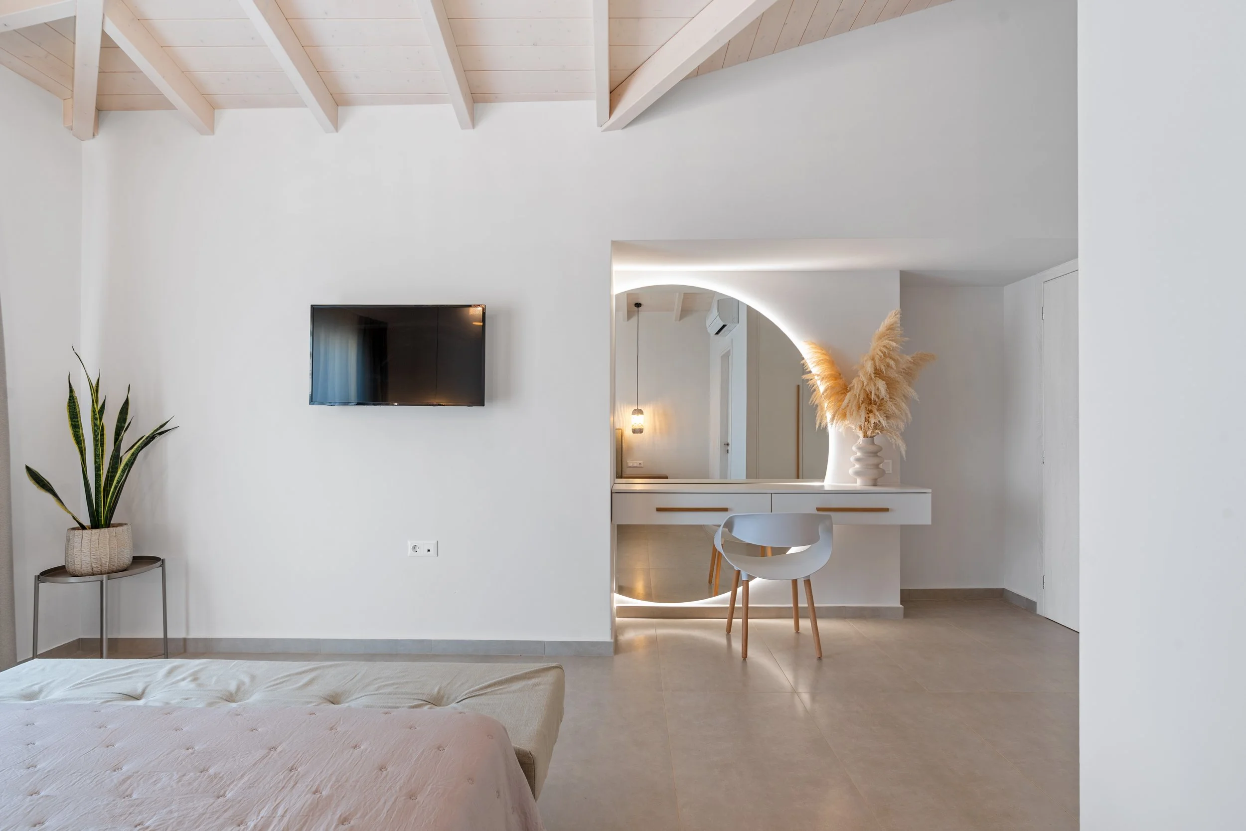 Minimalist bedroom with white walls, a wall-mounted TV, a potted plant on a side table, a bed with beige bedding, and a vanity area with a large mirror, chair, and decorative pampas grass.