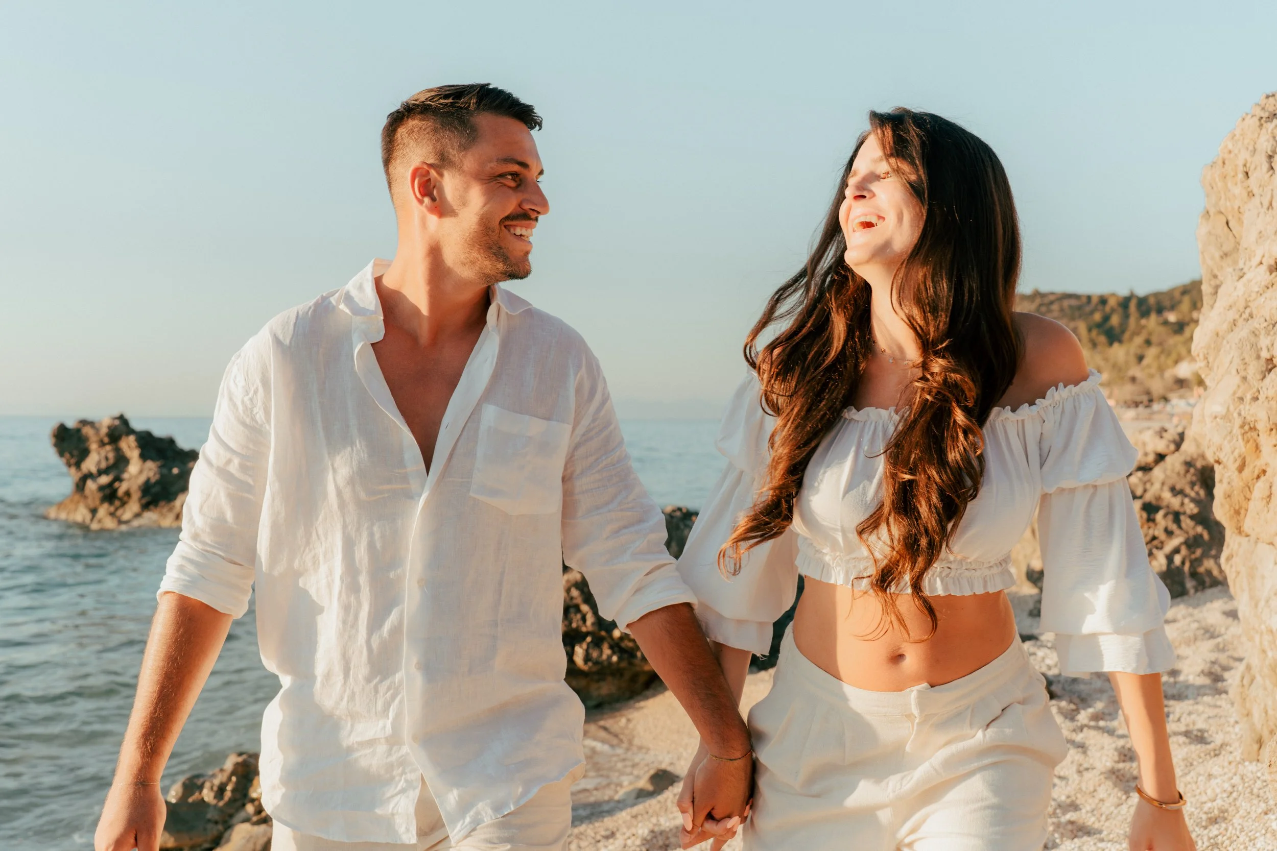 A happy couple holding hands and walking along a rocky beach at sunset, smiling and enjoying each other's company.