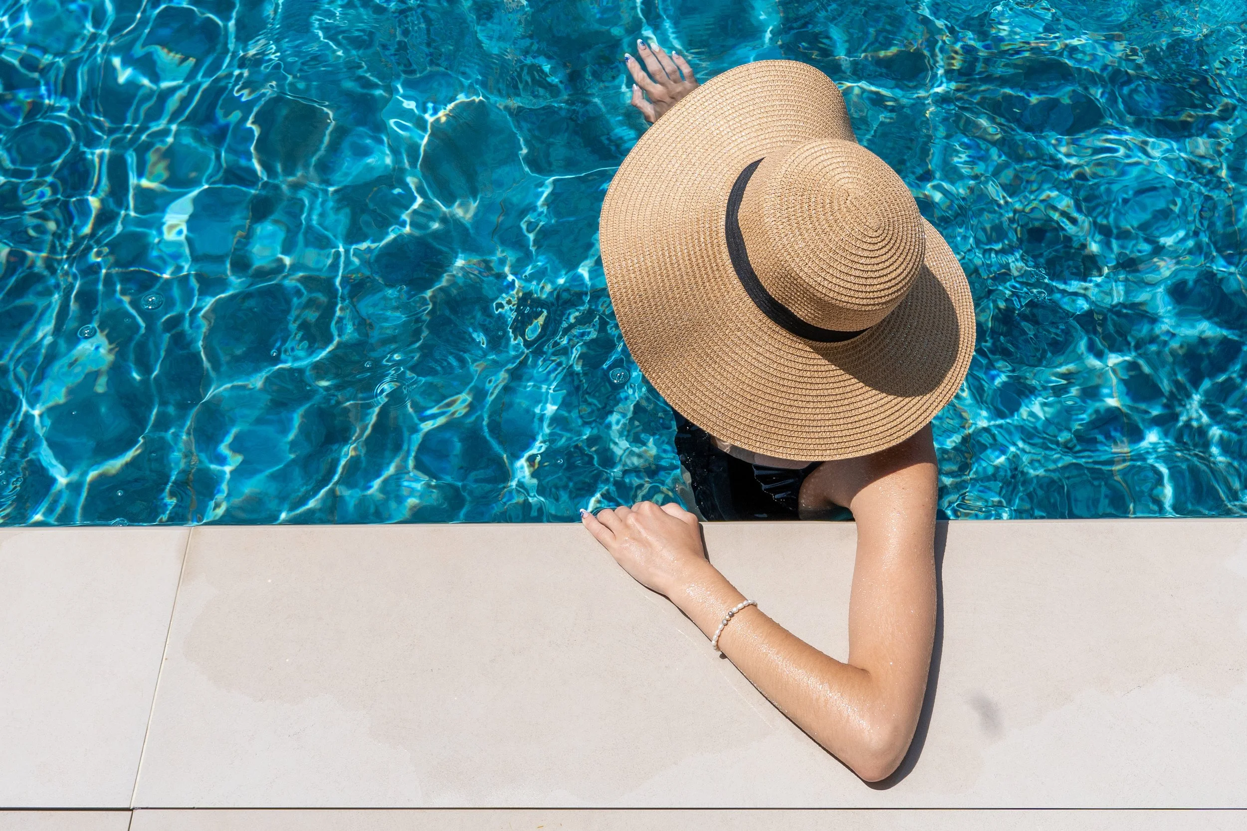 A woman with a large straw sunhat and a pearl bracelet relaxing at the edge of a swimming pool.