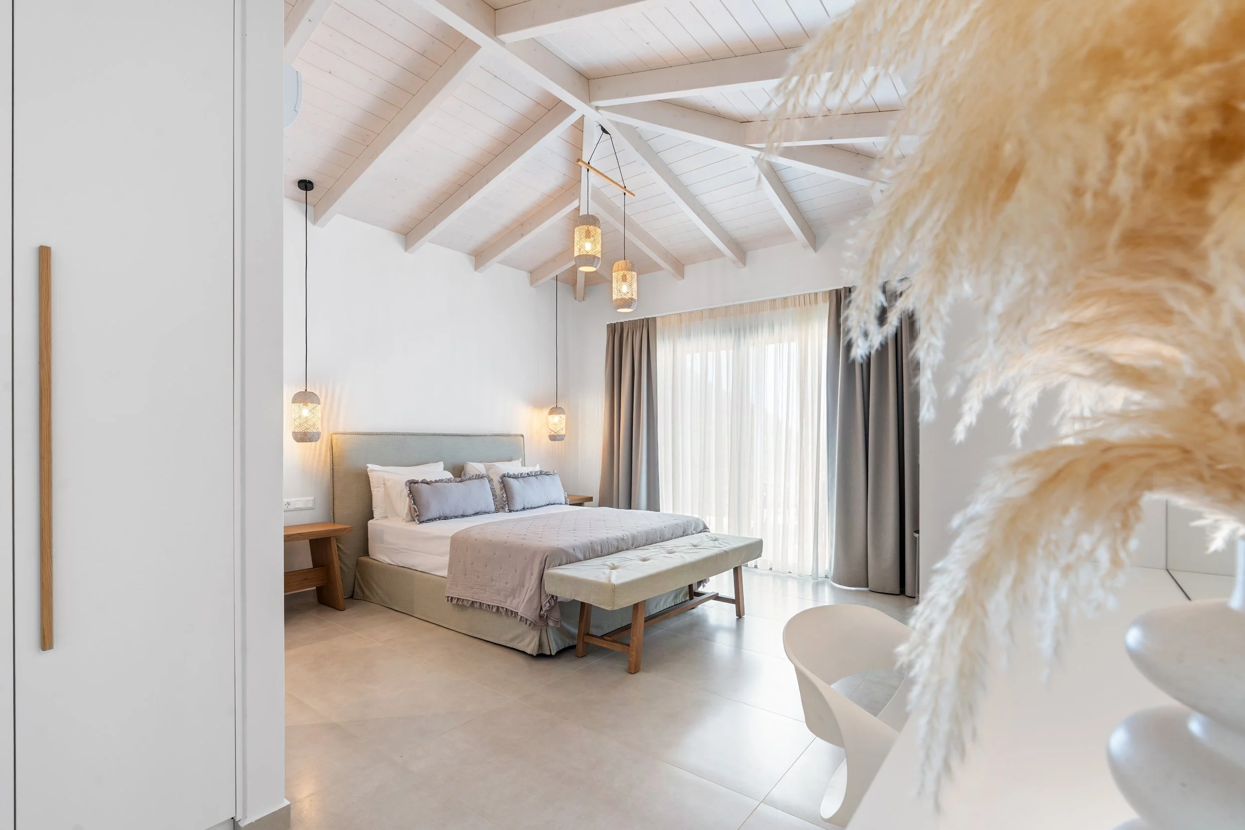 A bedroom with a vaulted ceiling with white wood beams, a large bed with light-colored linens and pillows, bedside tables with hanging pendant lamps, large windows with sheer curtains, and a bench at the foot of the bed, with a piece of decorative pa