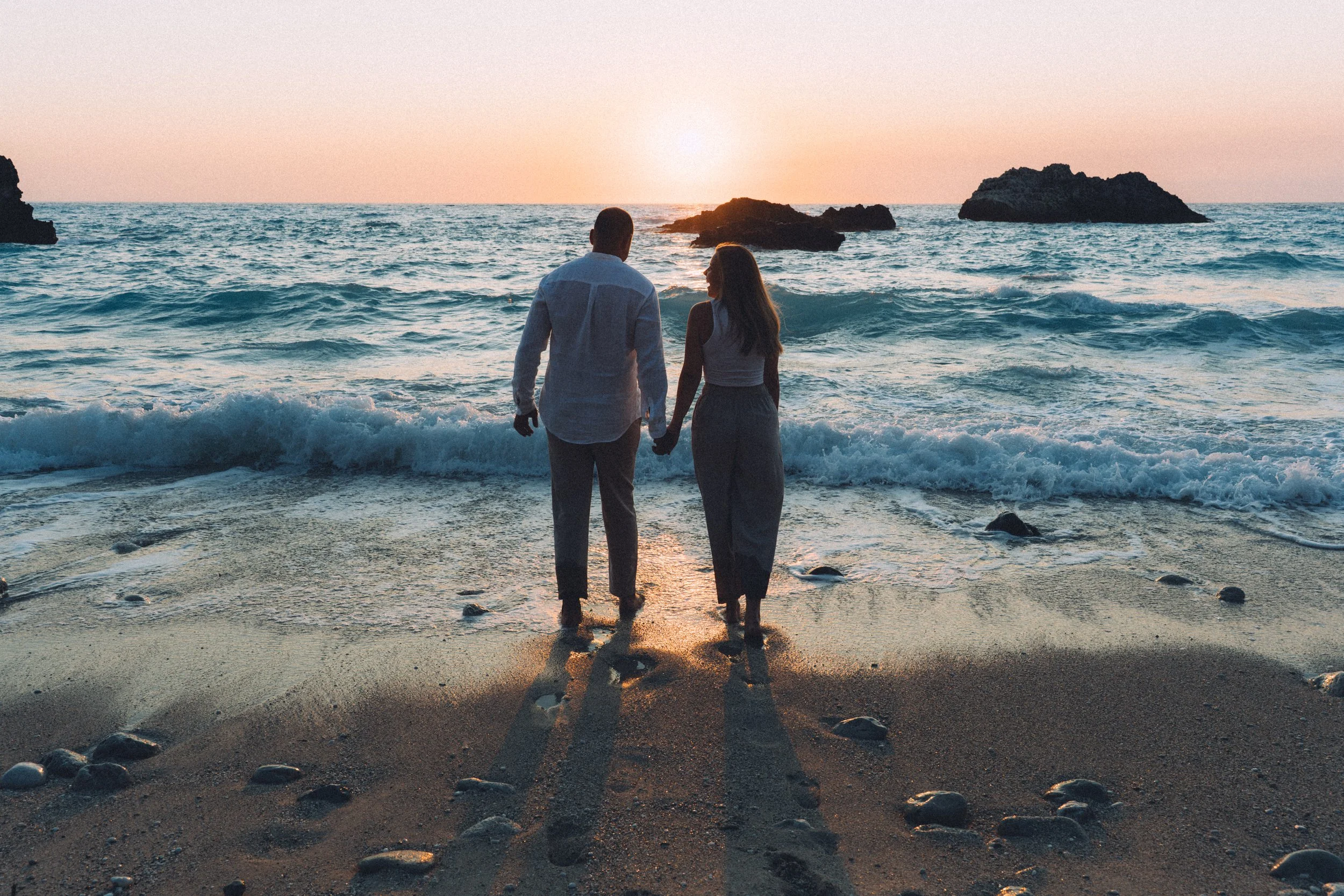 A couple holding hands at the beach during sunset, facing the ocean with rocks in the water.