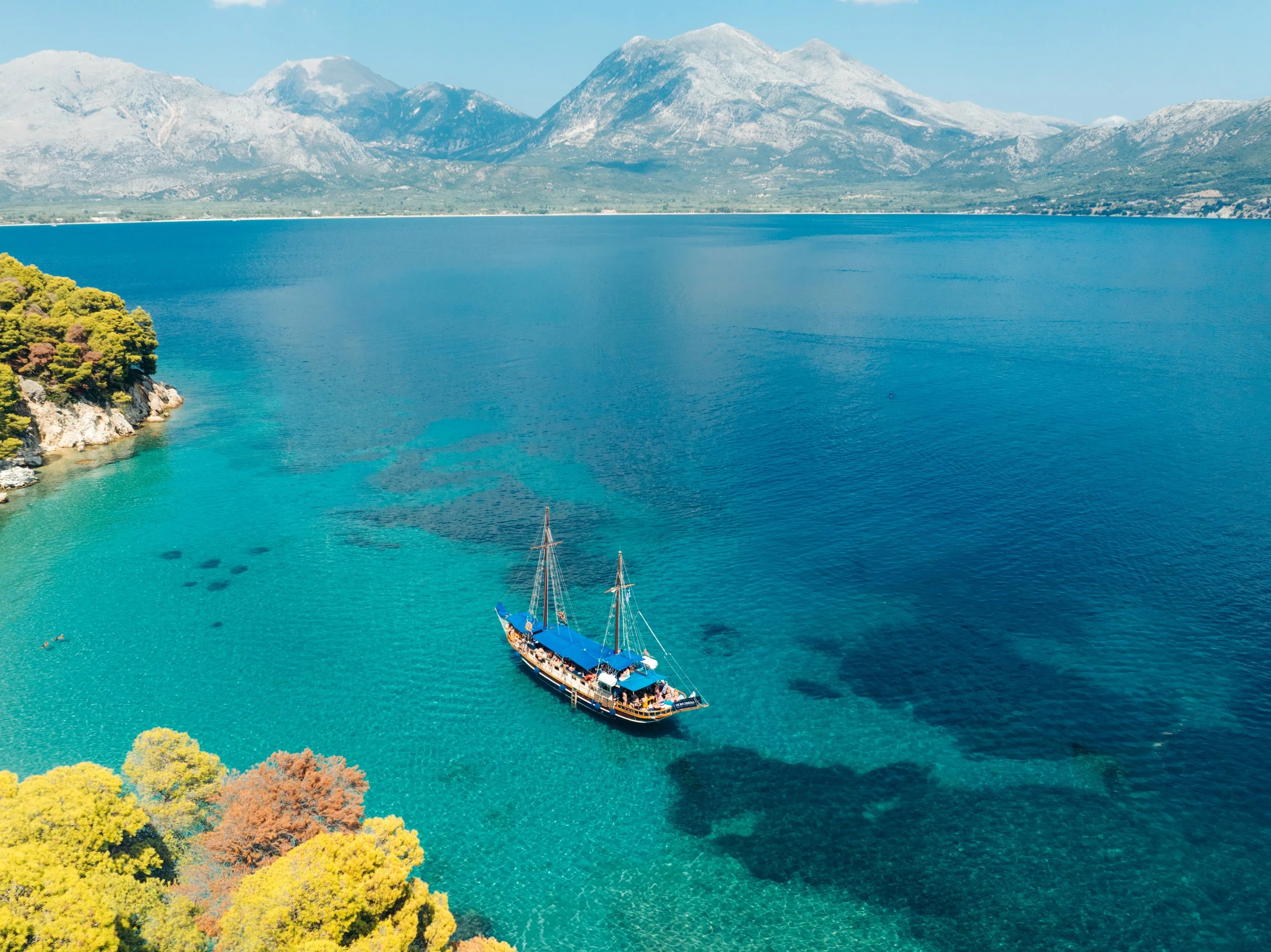 A sailboat with a blue canopy floats on clear turquoise water near lush green and yellow trees with a mountainous landscape in the background.